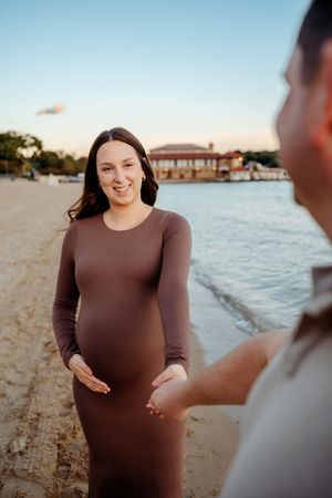 A person in a brown dress smiles during a beachside maternity photoshoot at sunset.