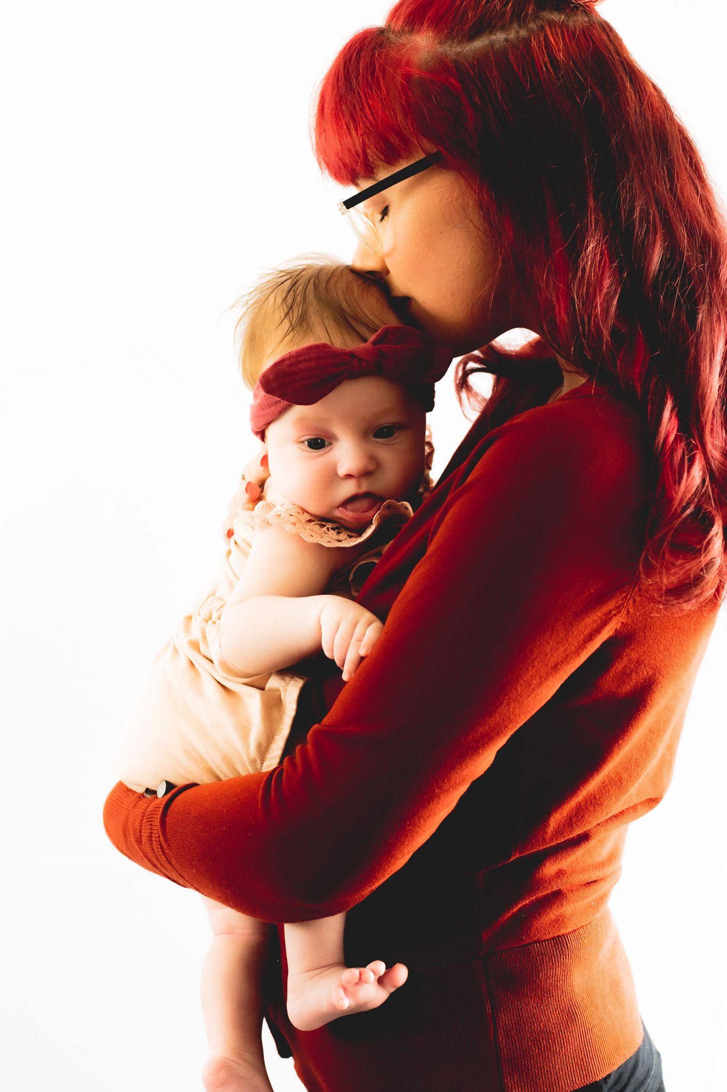Parent with bright red hair holds baby wearing matching red bow in warm lighting.