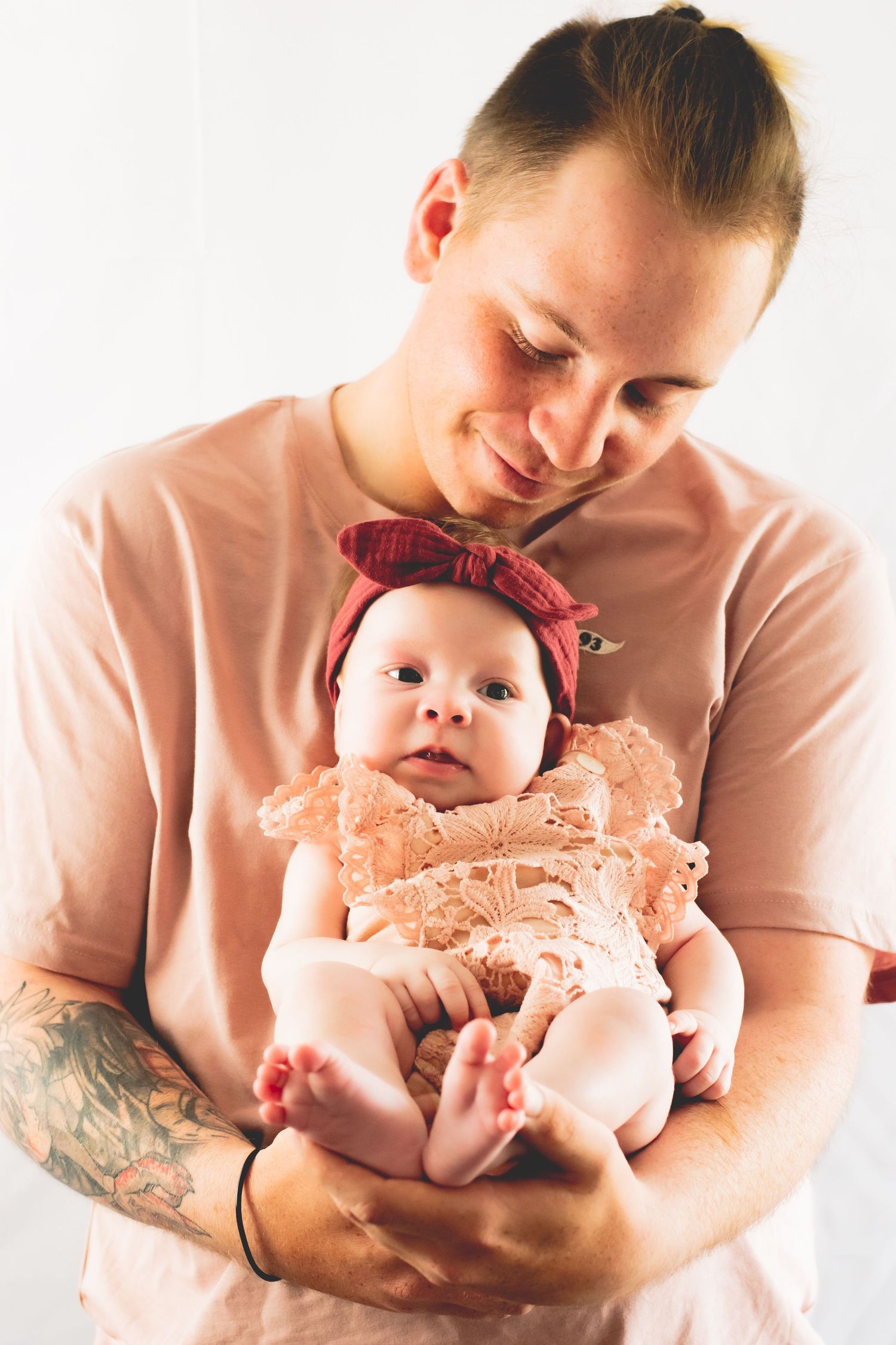 Parent in pink shirt holds baby wearing lace outfit and red bow headband.