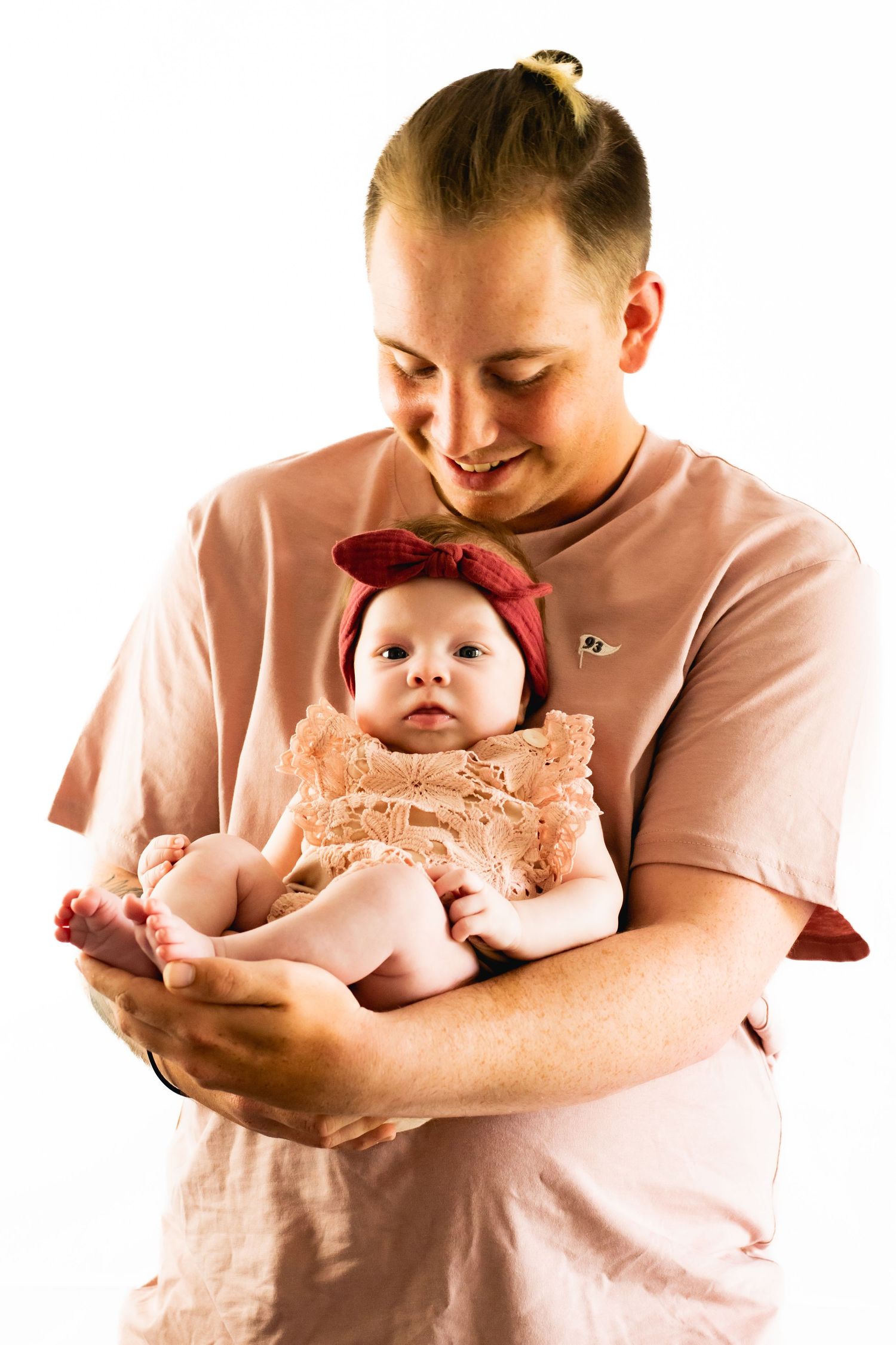 A person in pink holds an infant wearing a peach dress and red headband.