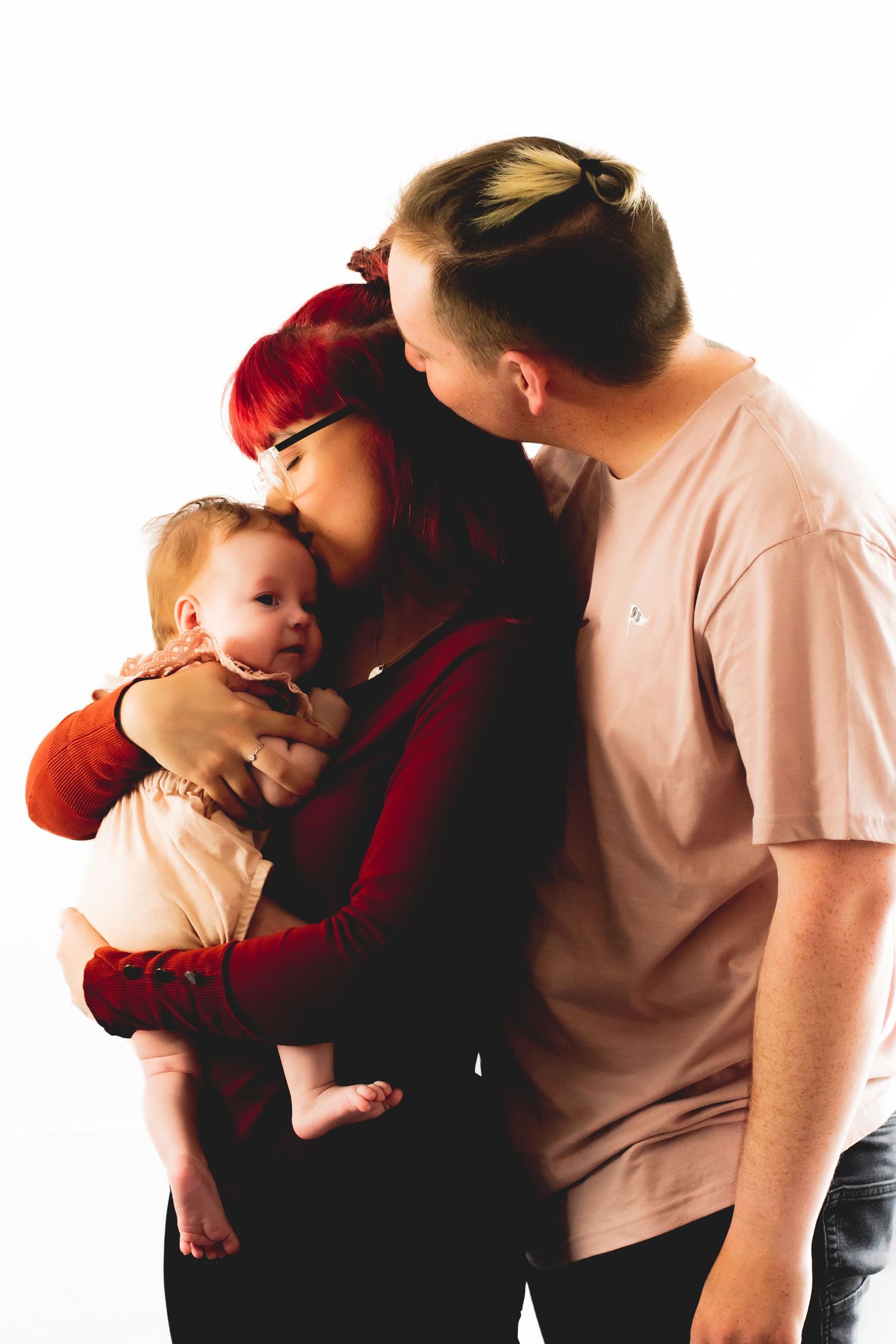 Family portrait showing parents in pink and red embracing their small baby.