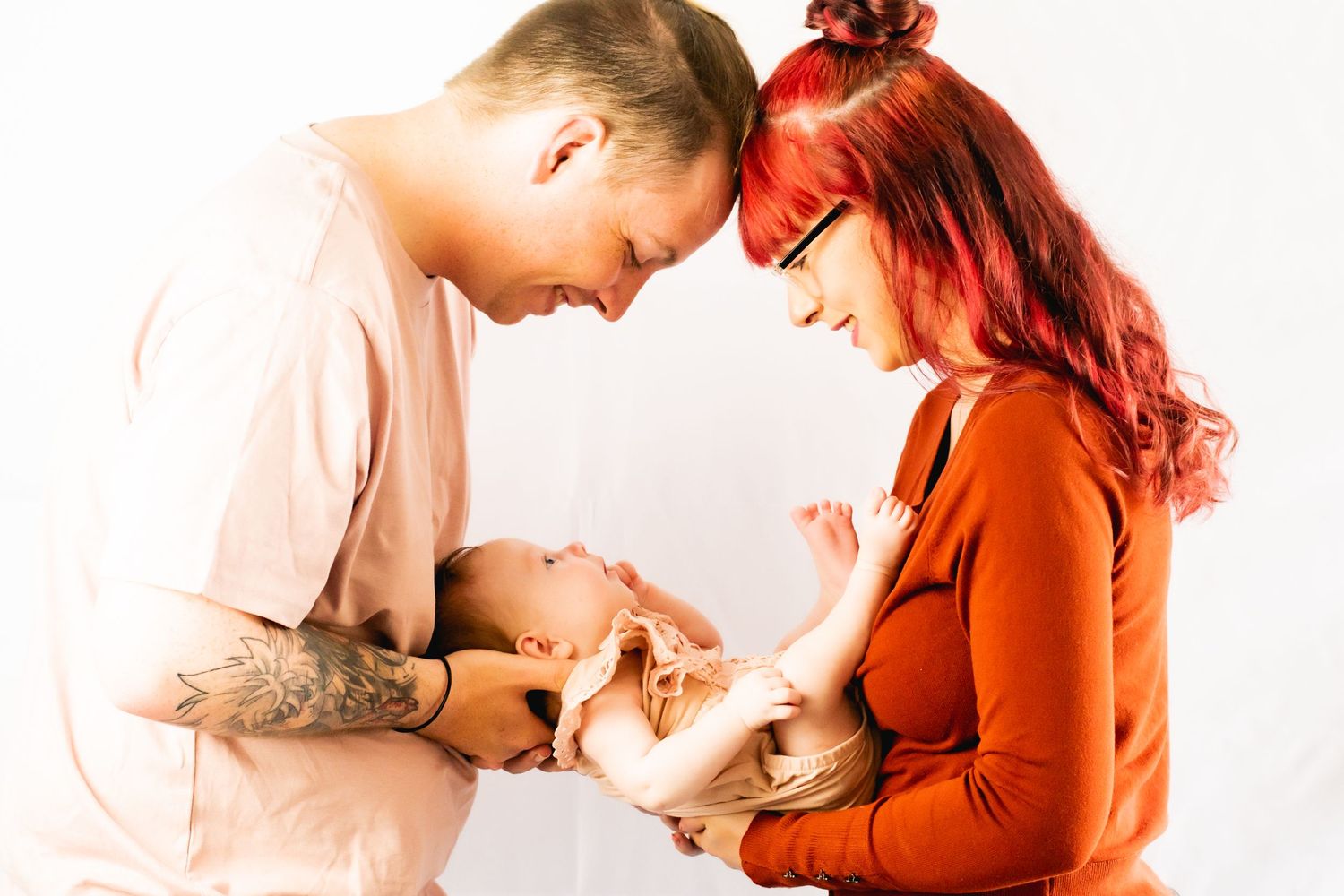 Intimate portrait of couple in pink and rust colored clothing gazing down lovingly at their infant.