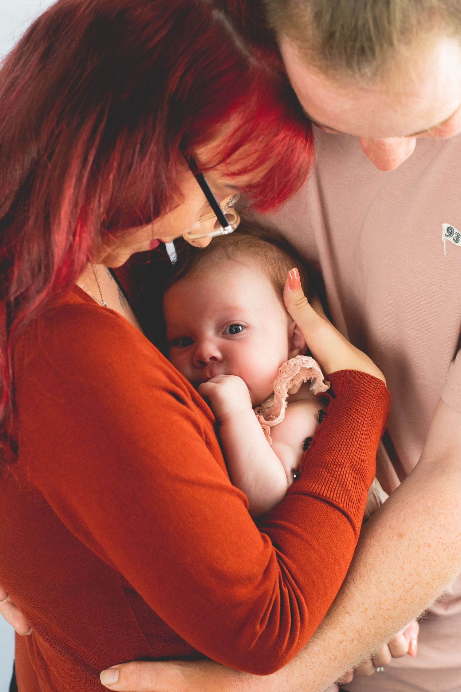 A tender family moment as parents cradle their baby while wearing red and beige clothing against a white background.