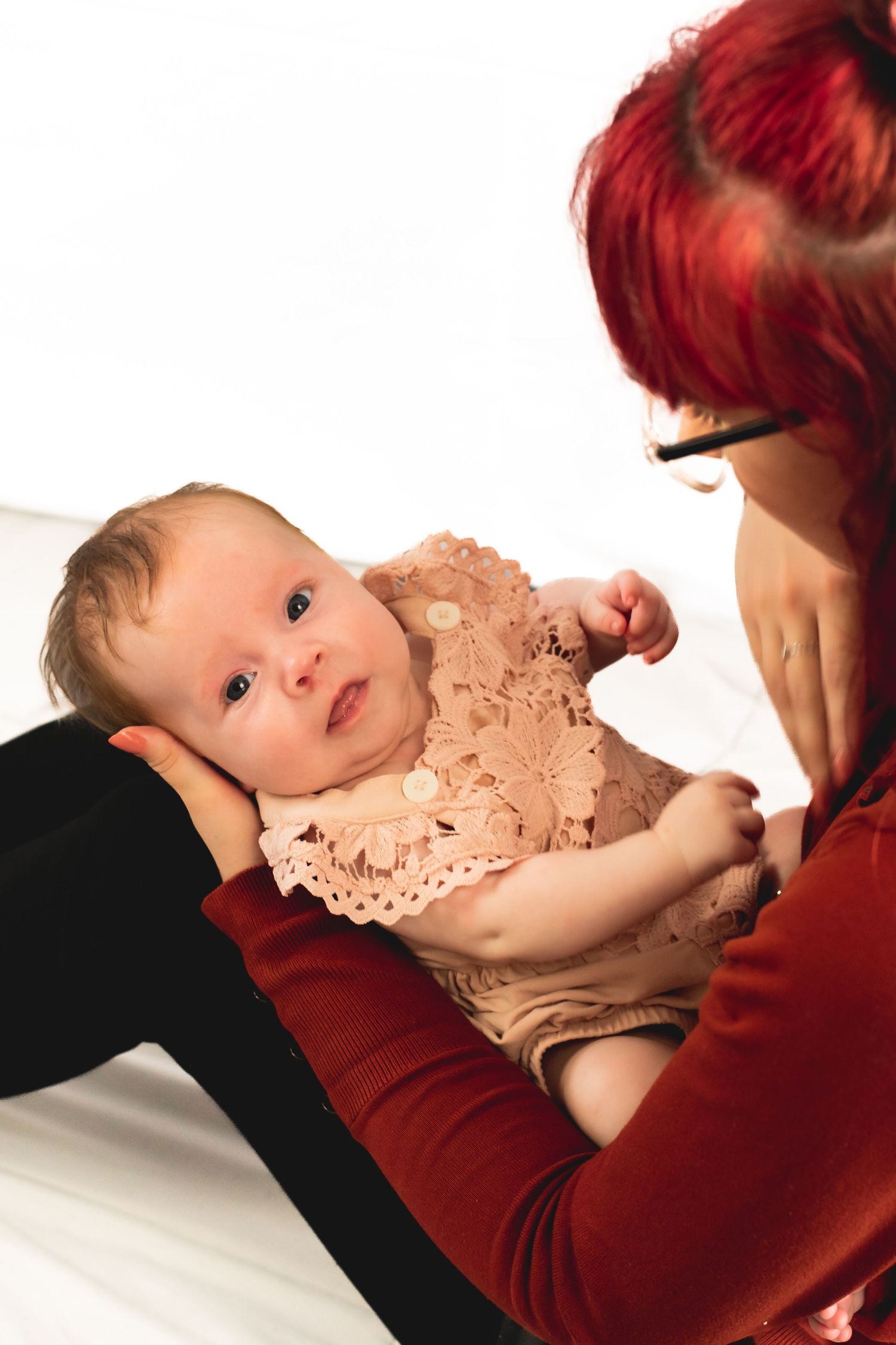 An infant in peachy lace outfit gazes up while lying on white surface.