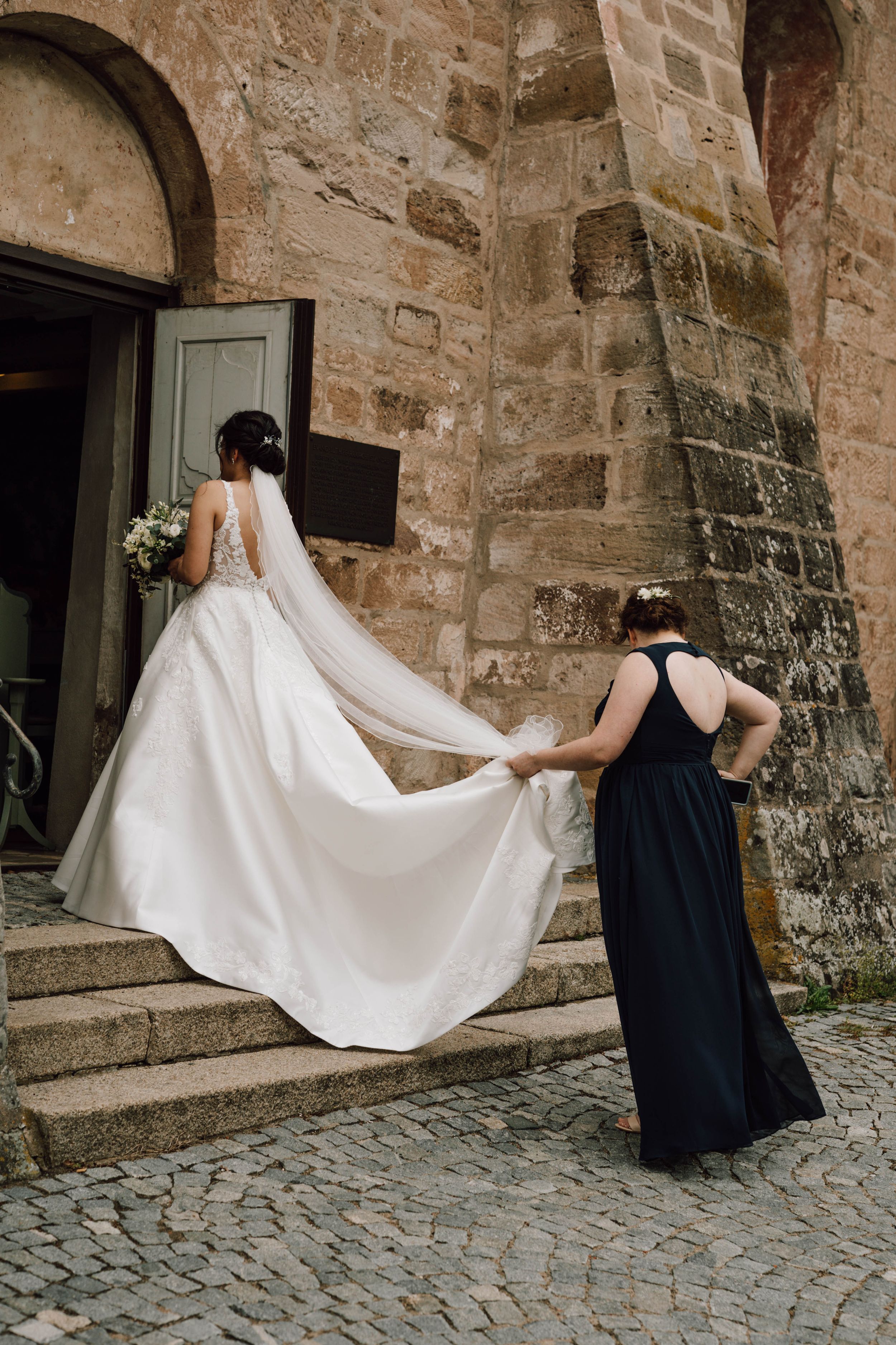 A flowing white wedding dress train being arranged on stone church steps.