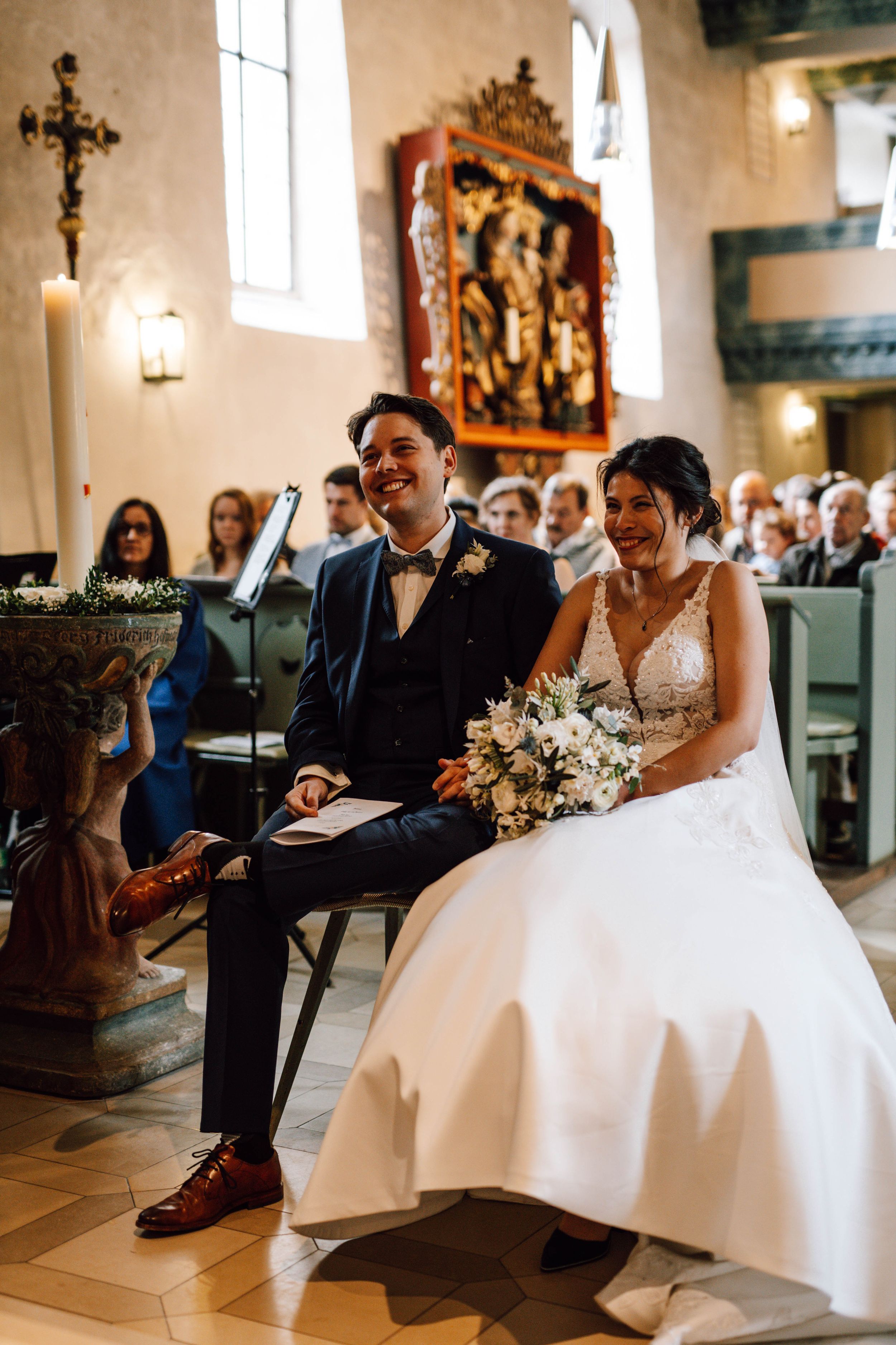 A couple shares a joyful moment during their church wedding ceremony, seated together in ornate wooden pews.
