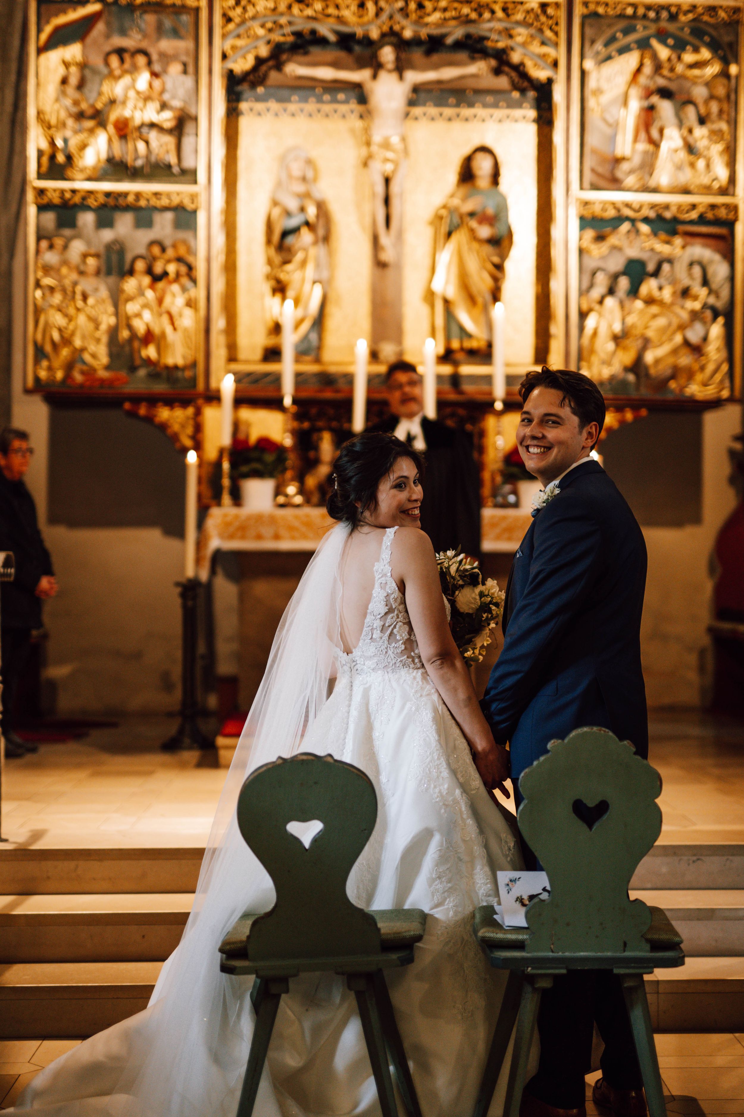 Couple sits on decorative chairs in a church with ornate golden religious artwork in the background.
