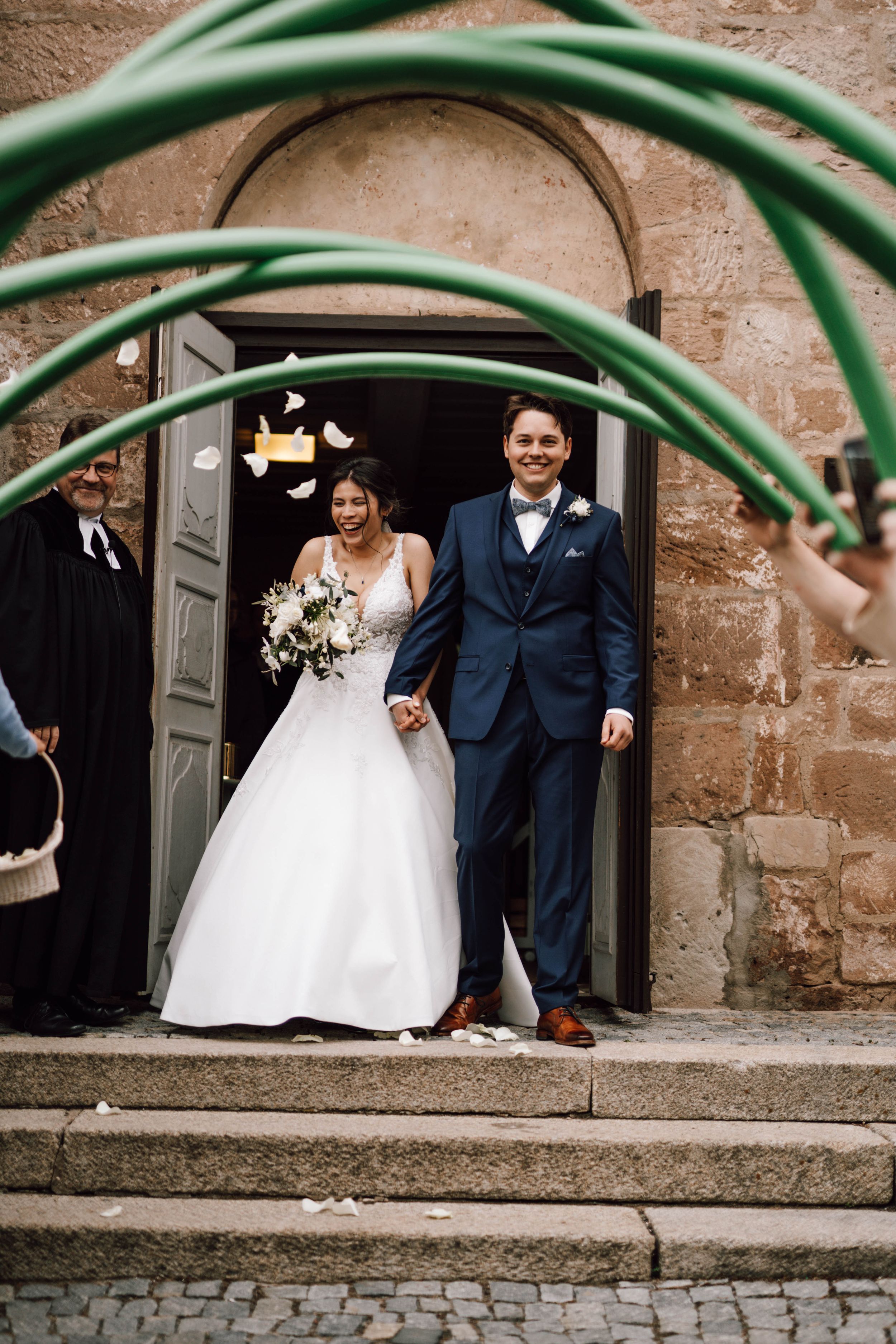 Newlyweds emerge from church doorway under green garden hose arch in playful wedding photo.
