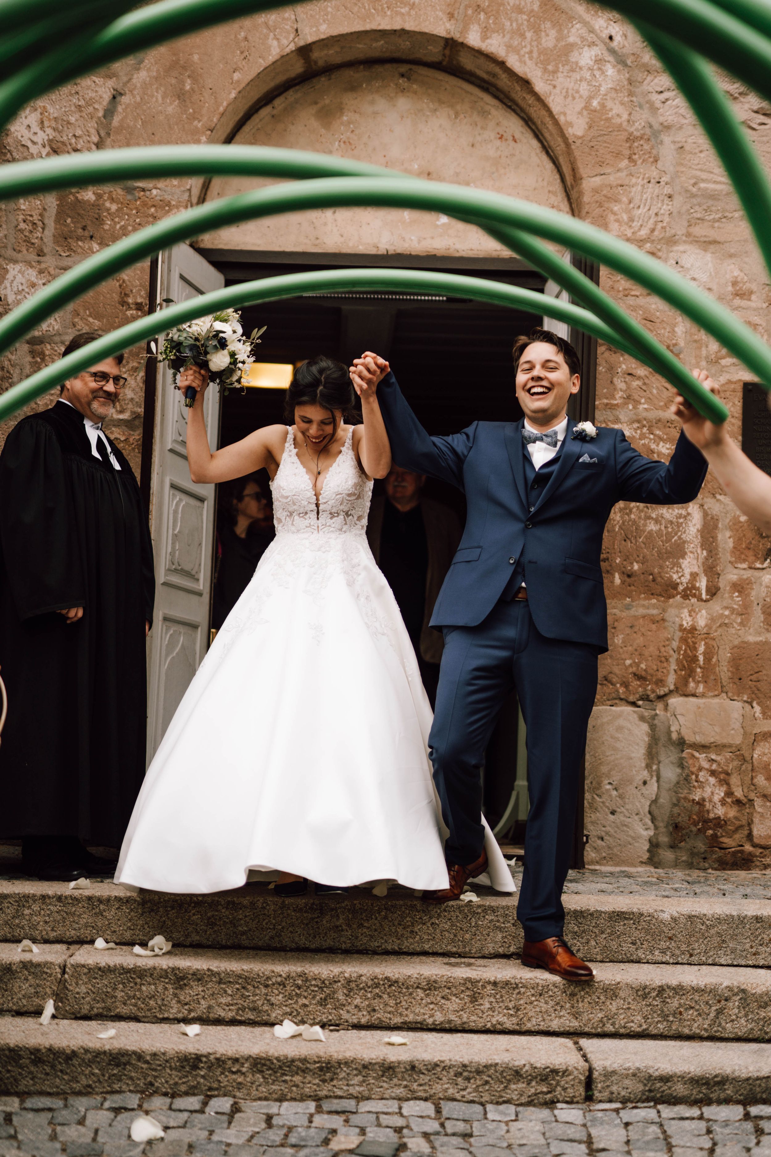 Happy couple celebrates under quirky green garden hose arch outside historic church building.