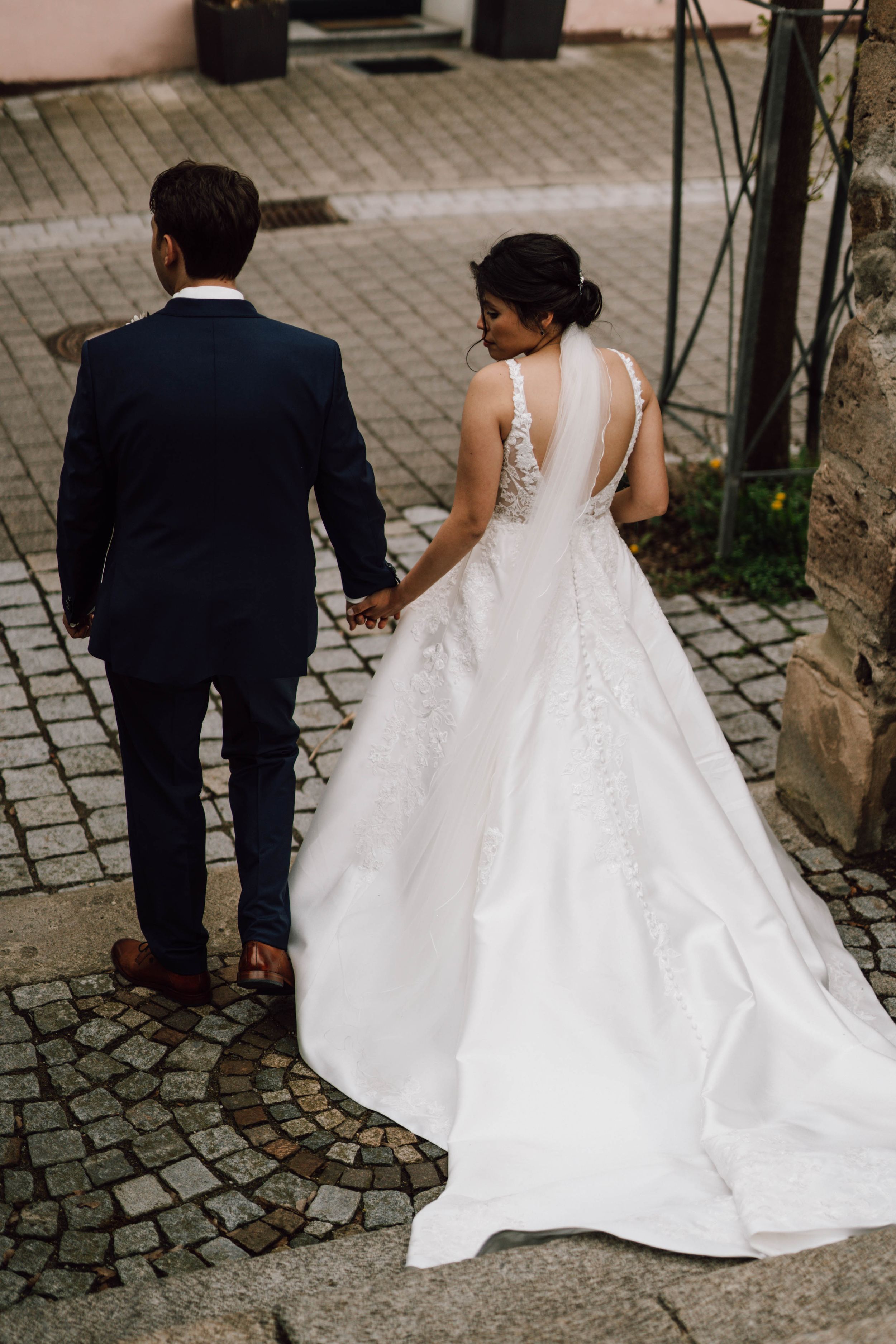 A couple in wedding attire walk along a cobblestone path, with the bride's elegant white dress trailing behind.