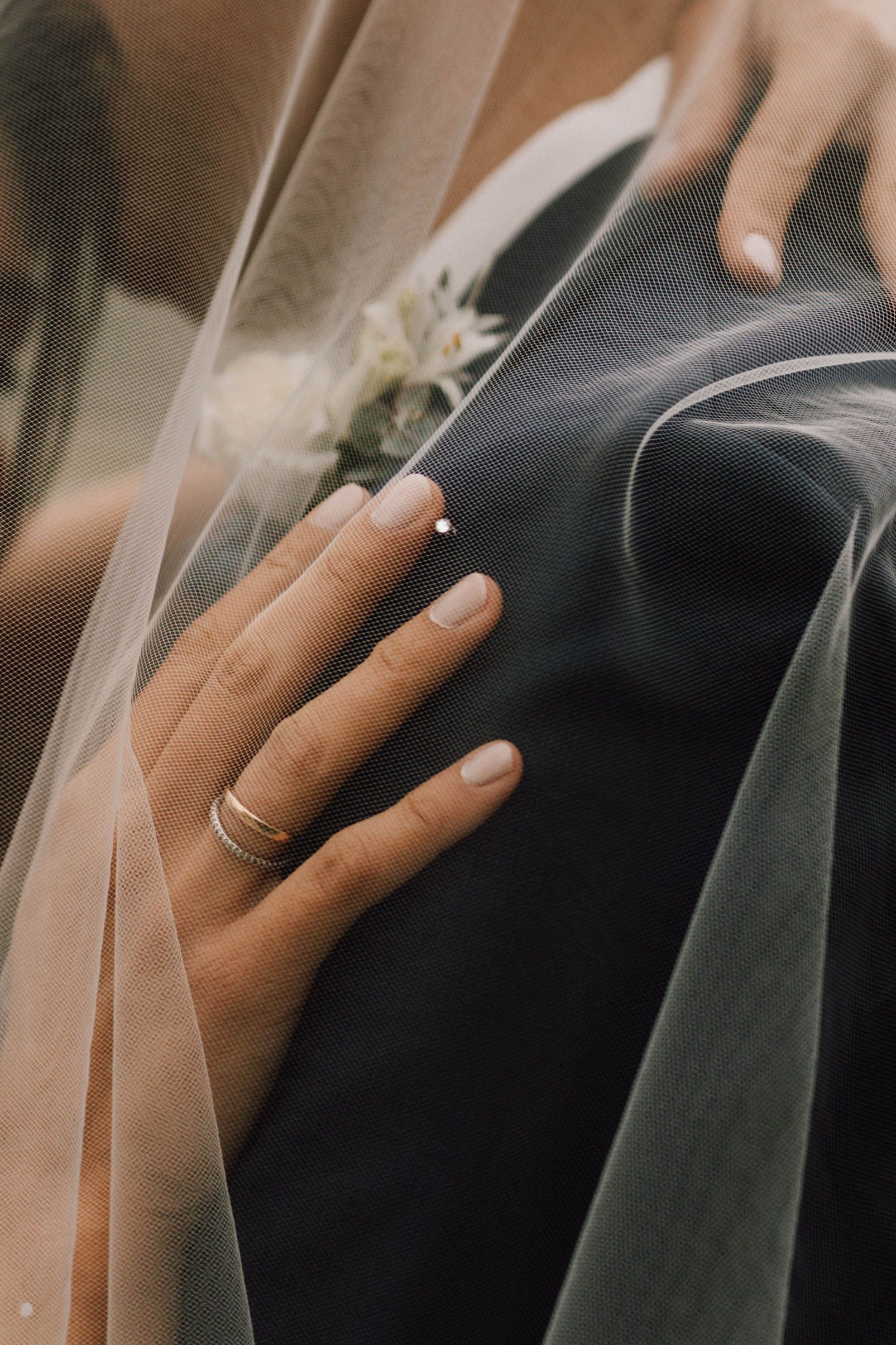 A close-up of a wedding ring and veil against a dark suit fabric.