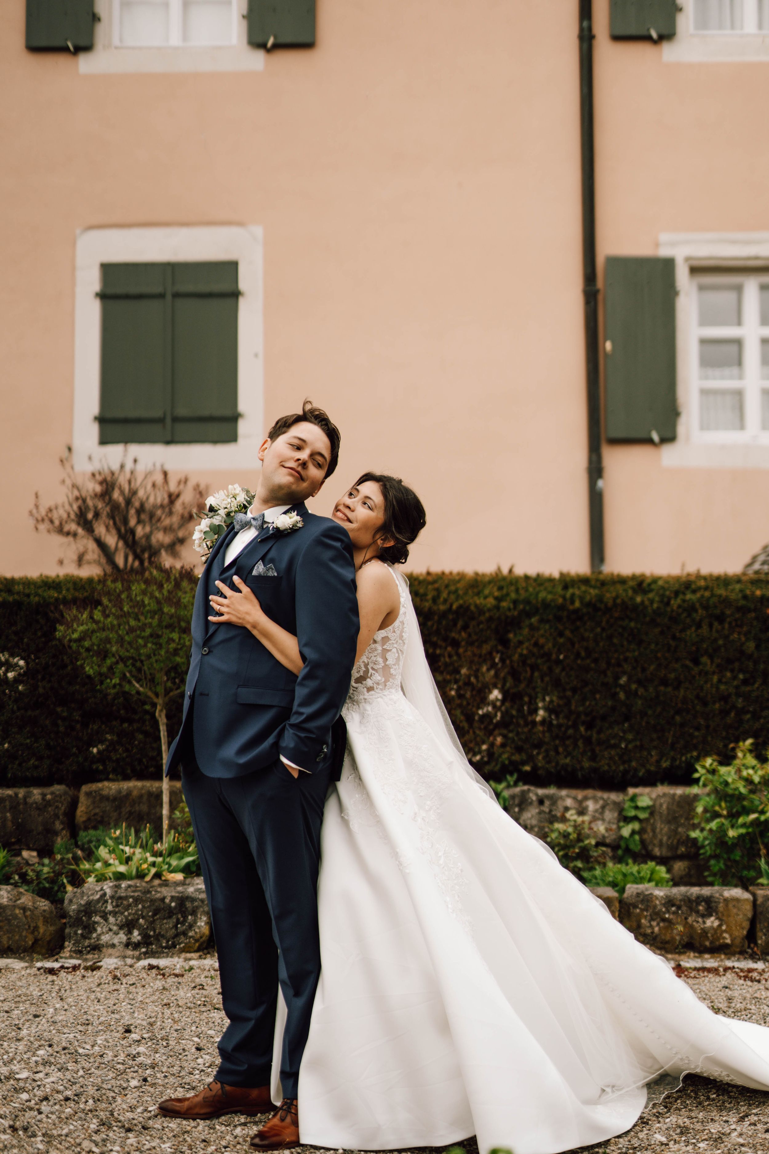Romantic wedding couple poses playfully against a peach-colored building with green shutters.