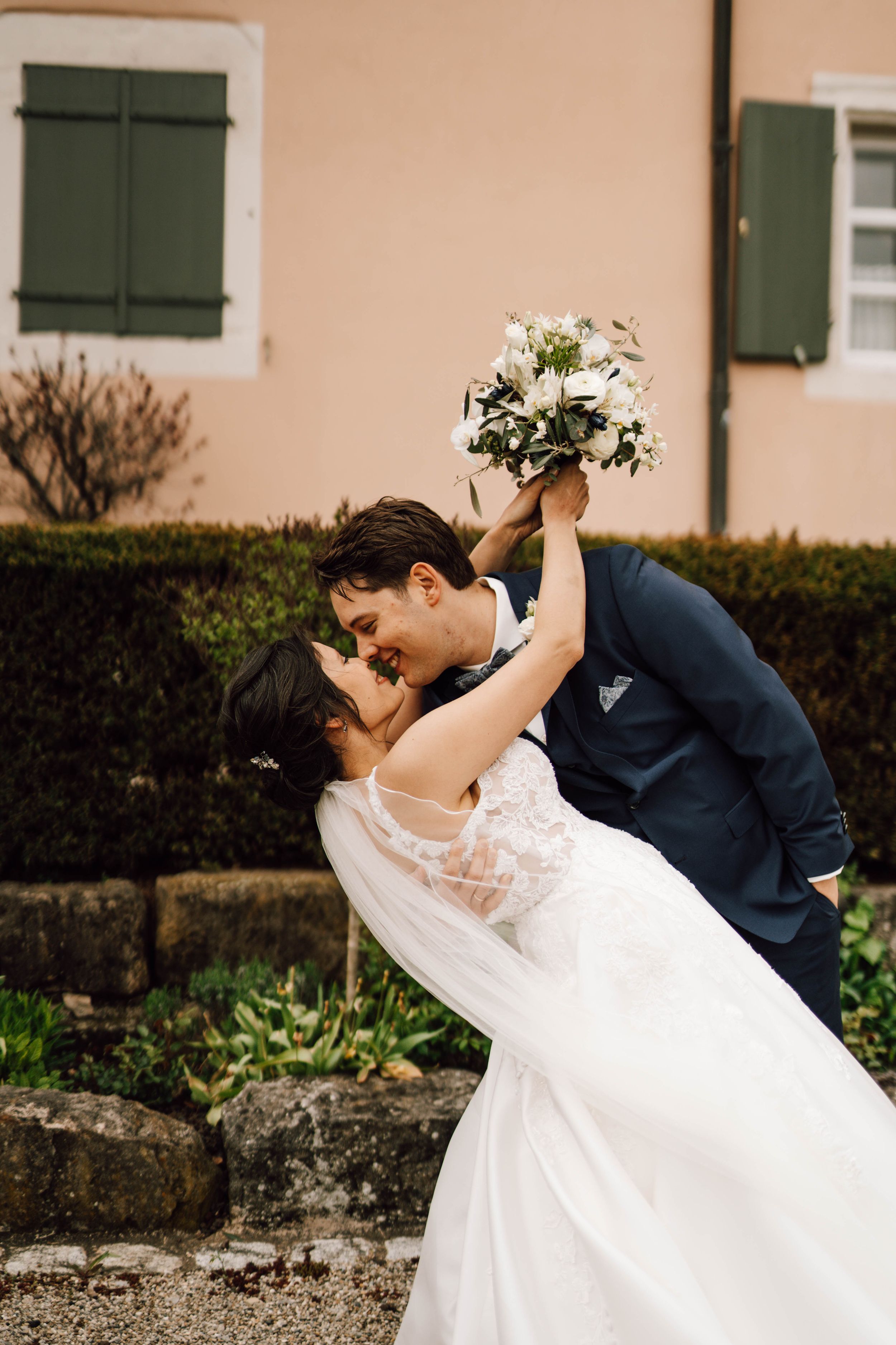 Wedding couple shares romantic dip pose in front of peach building with green shutters.