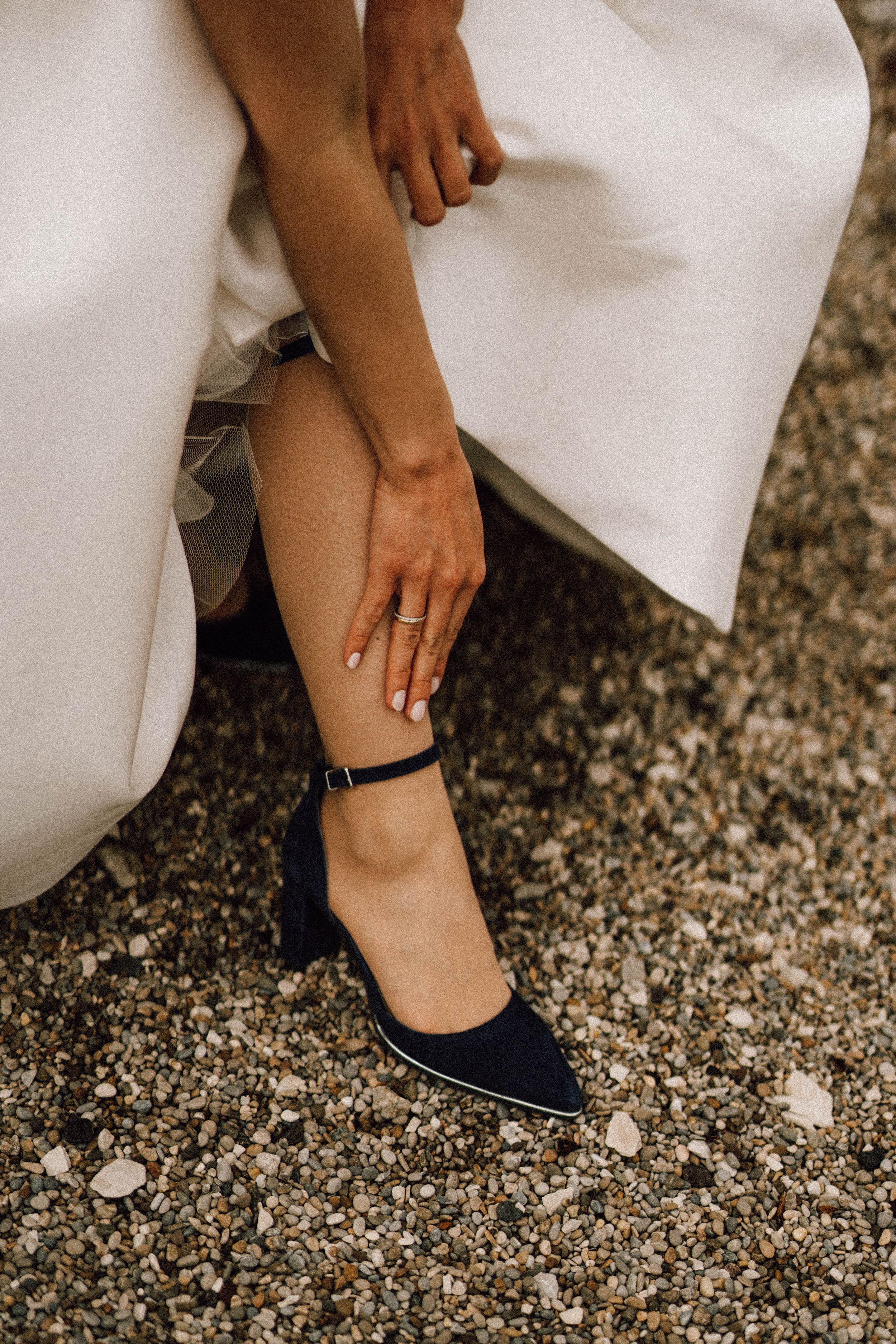 Close-up view of a navy blue flat shoe with ankle strap worn with a white dress on gravel ground.