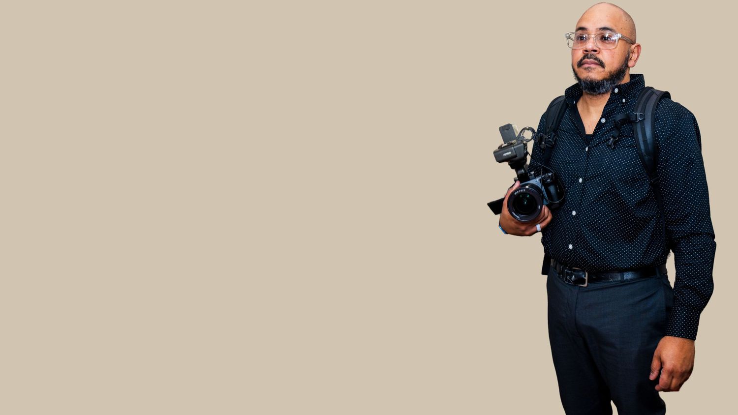 Professional photographer in navy blue outfit holding camera equipment against neutral background.