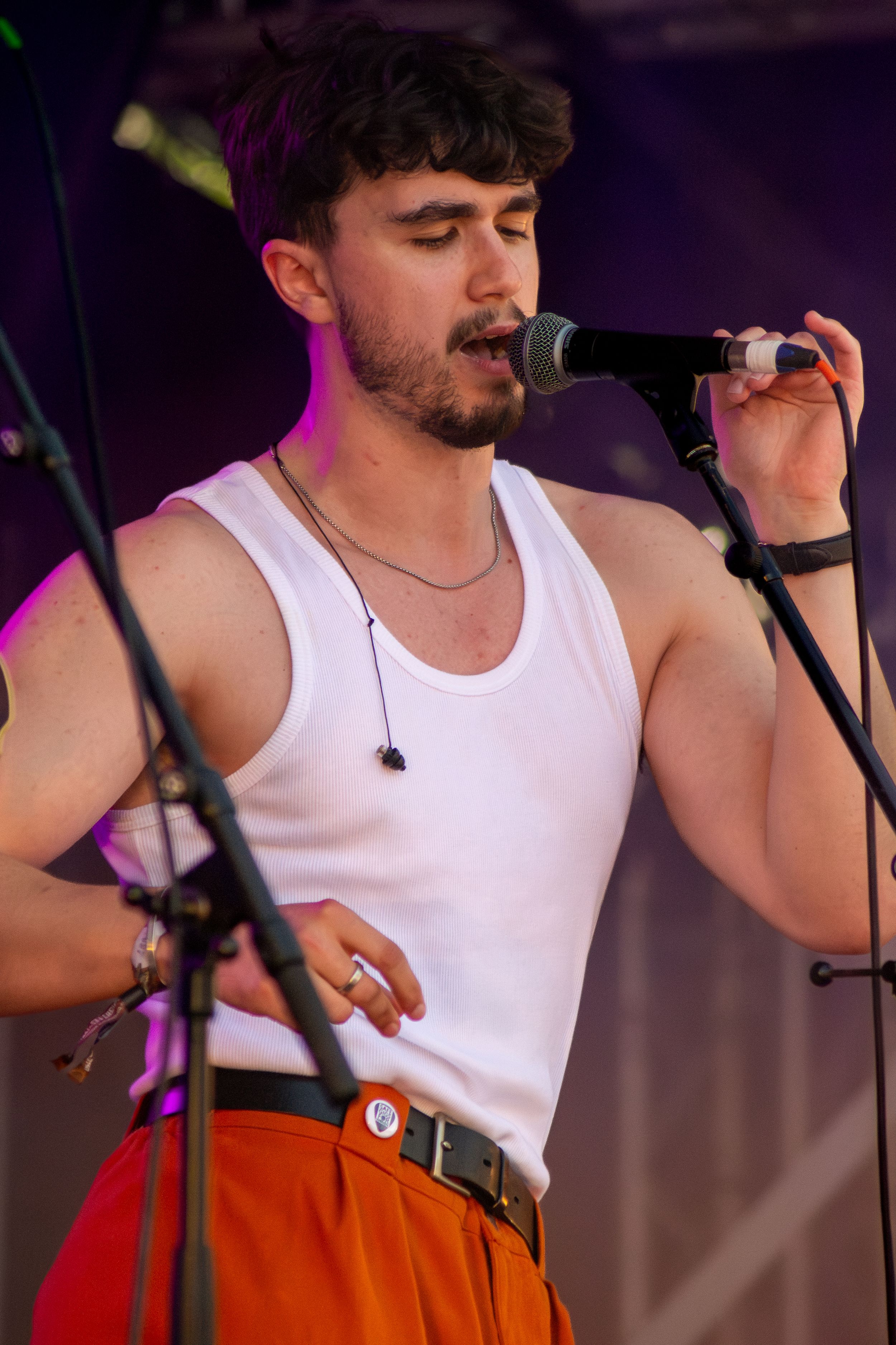 Performer in white tank top singing into microphone on stage with purple lighting.