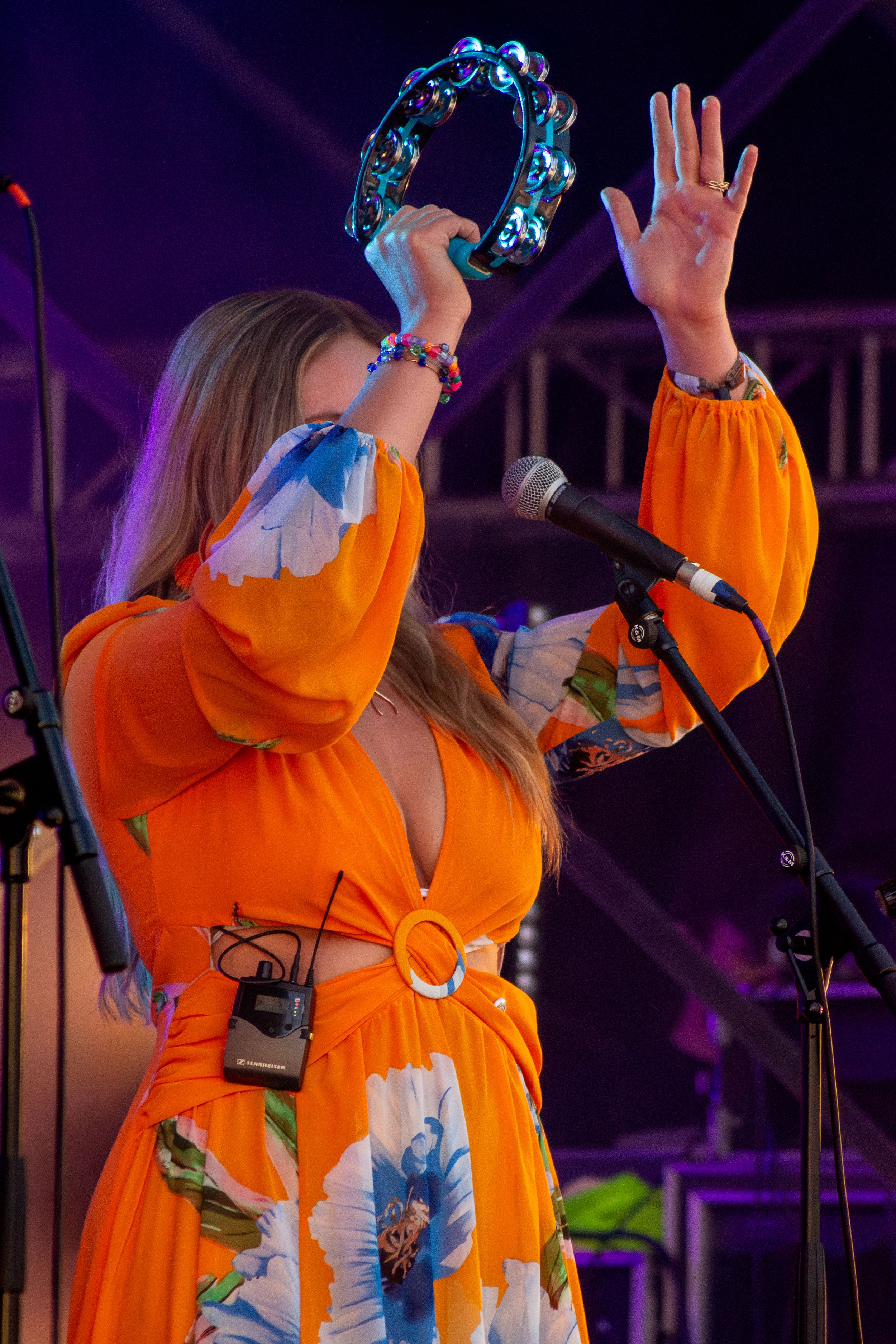 Performer in orange floral dress playing tambourine on stage with raised arms.