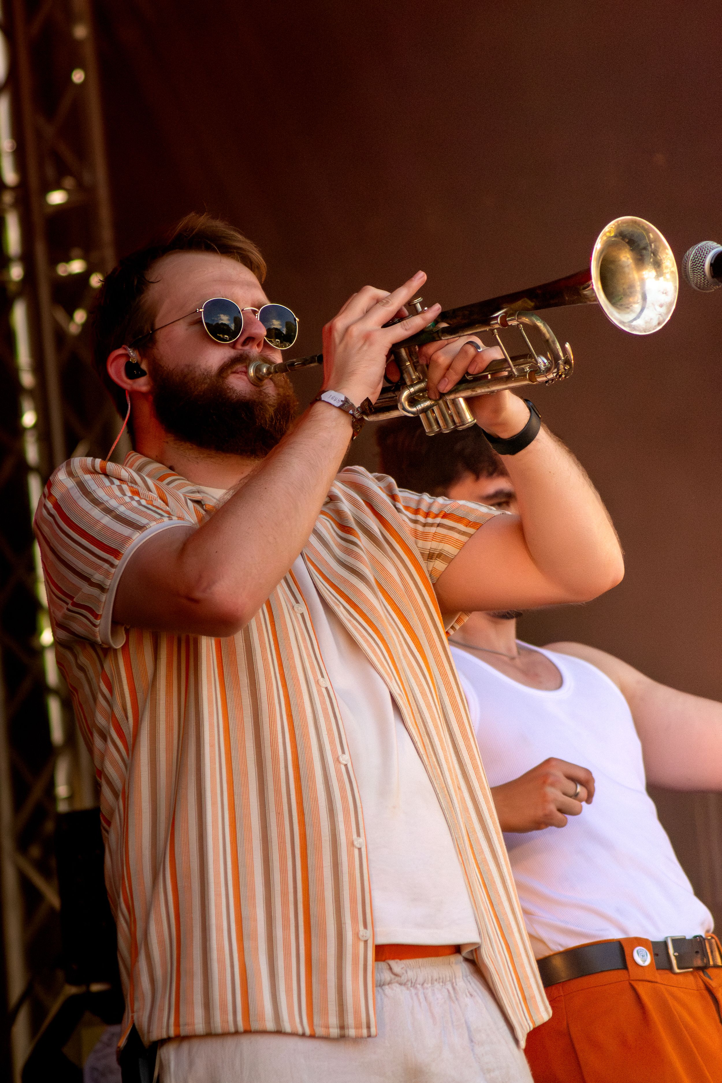 Musician playing trumpet on stage wearing striped shirt and sunglasses during live performance.