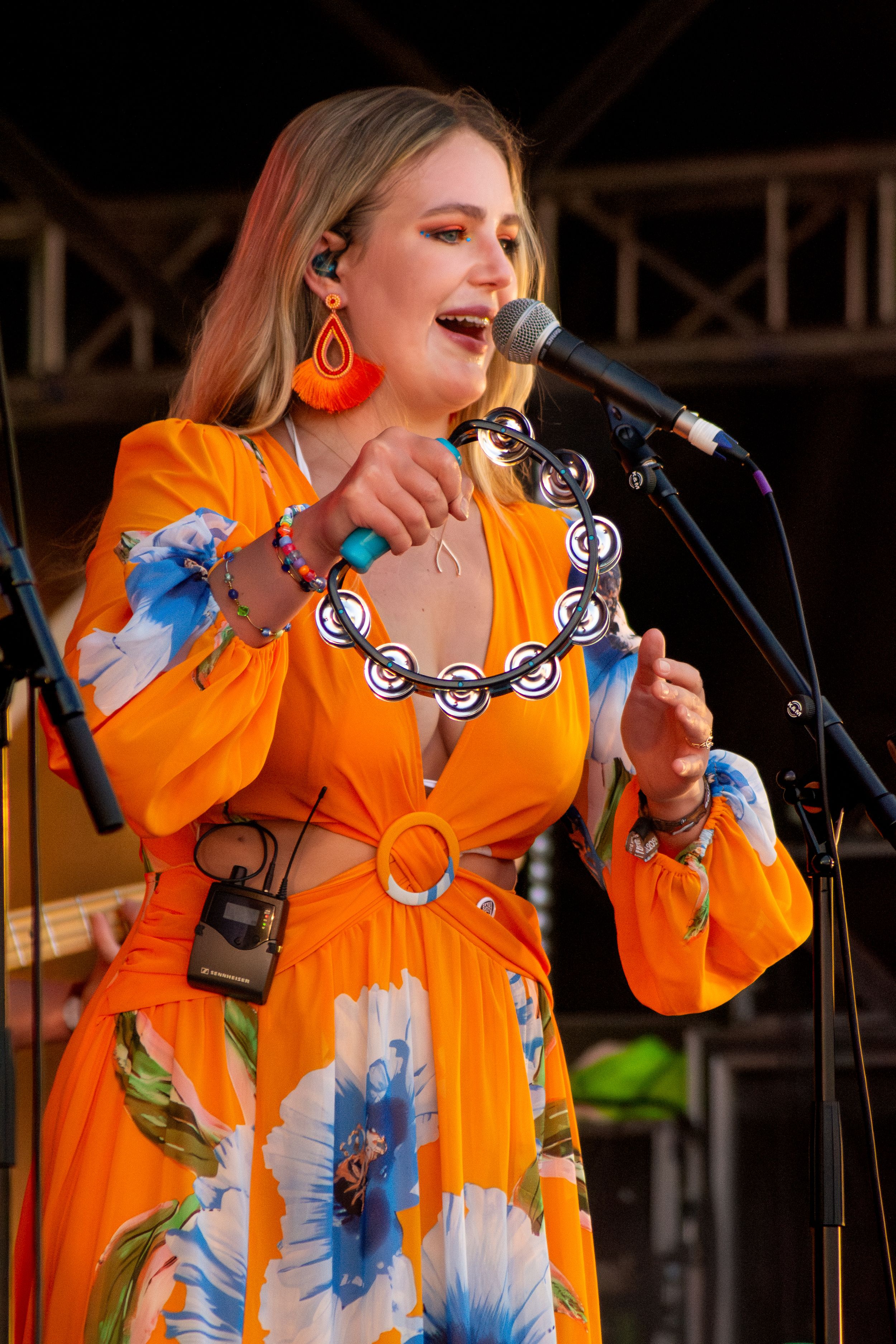 Singer in orange floral dress with statement necklace performing on outdoor stage.