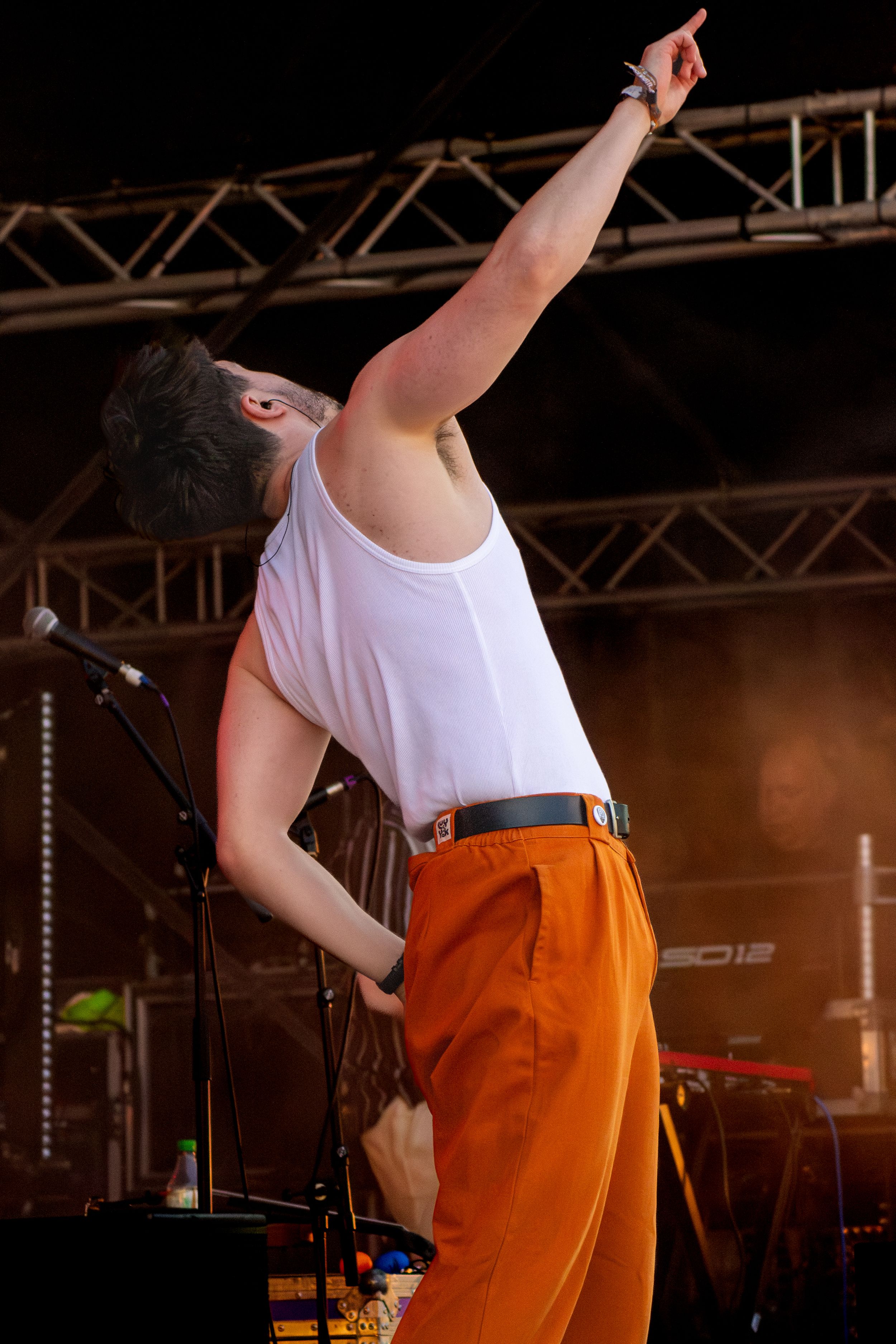Performer in white tank top and orange pants pointing upward dramatically on stage.