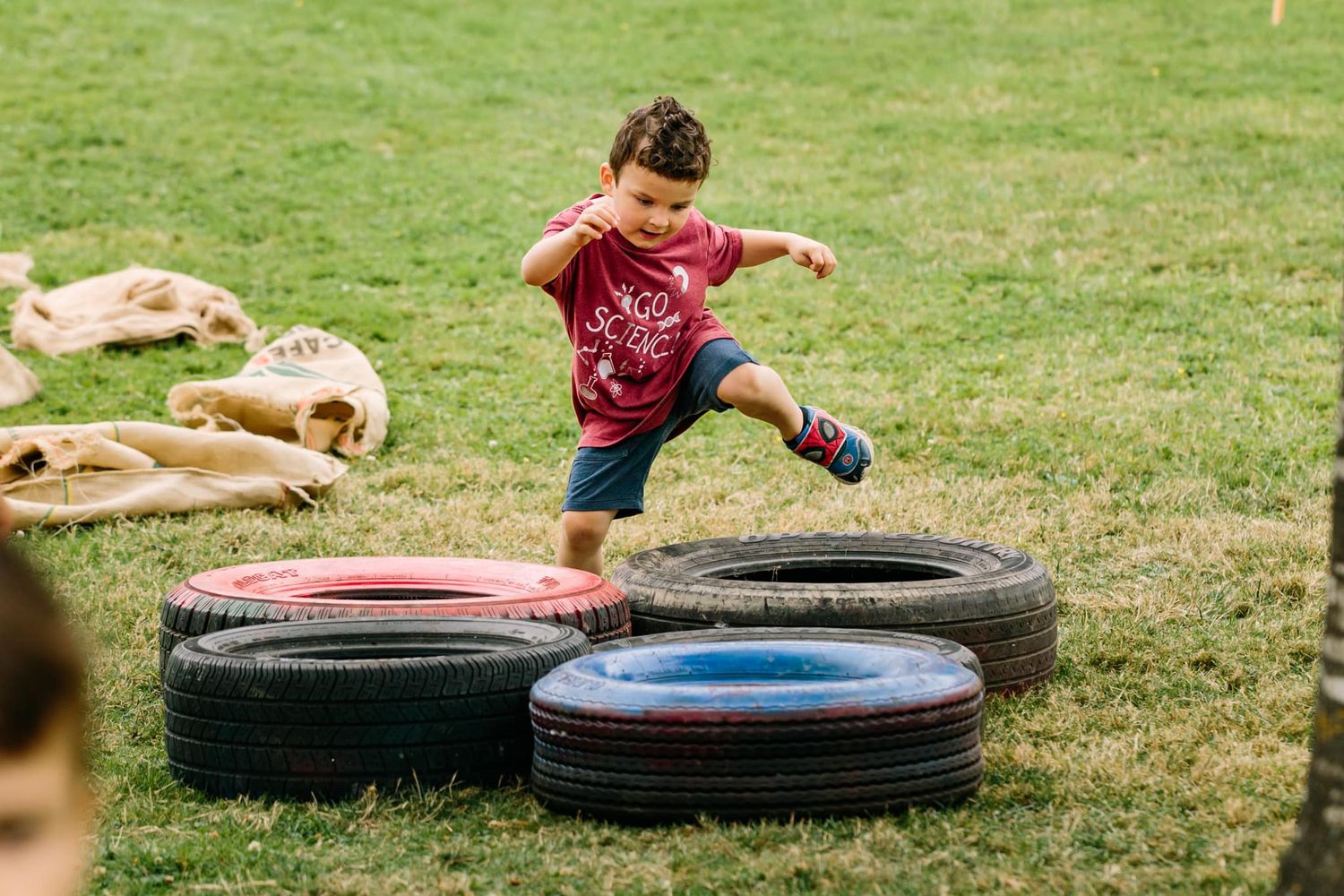 Child jumping and playing on tire obstacles in a grassy backyard.