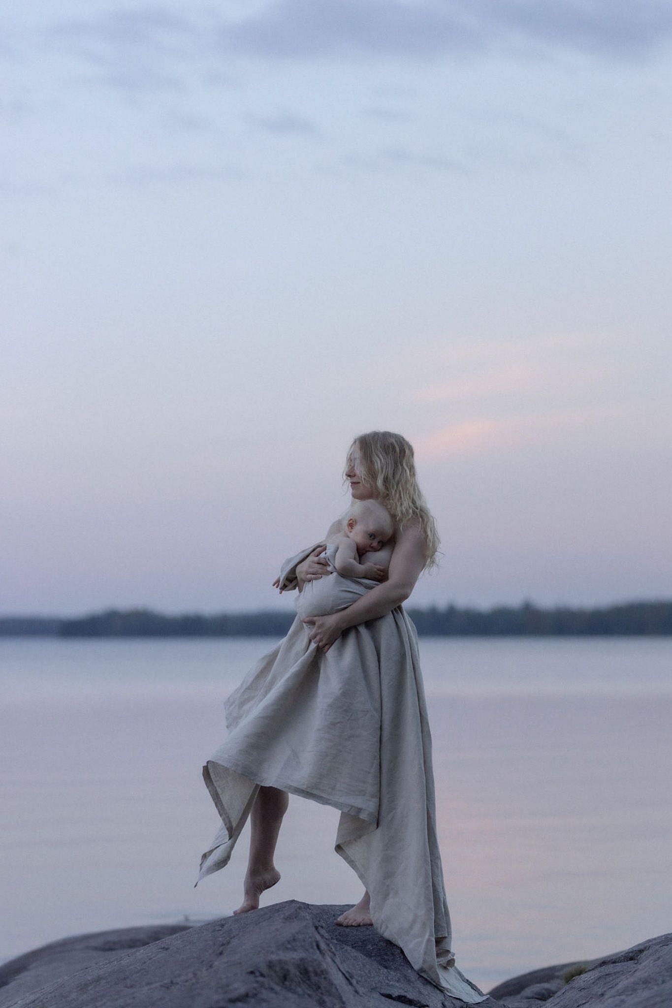 A mother in a flowing dress stands on rocks by a calm lake at dusk with a moody purple sky in the background.