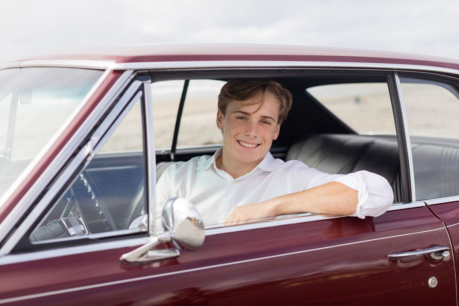 A smiling high school senior sits in a vintage maroon car with their arm resting on the window frame on a bright sunny day.