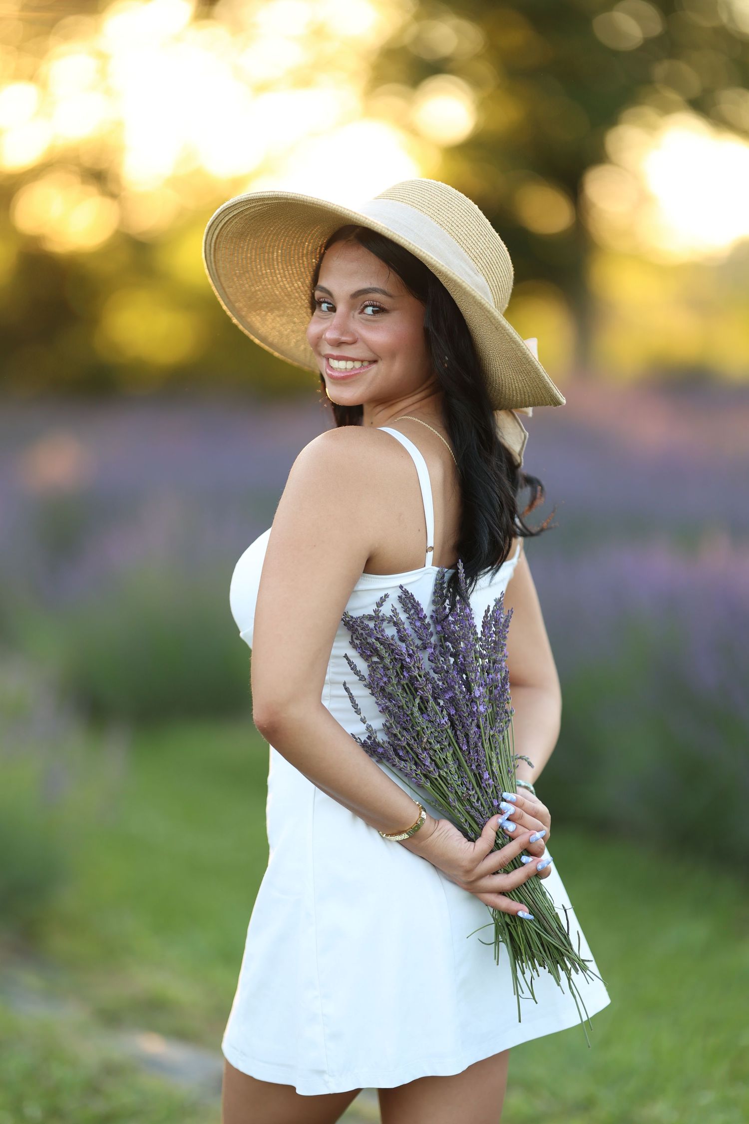 A senior girl in a white dress and sun hat holds fresh lavender flowers in a sunny field at golden hour.  Middle TN Senior Photographer