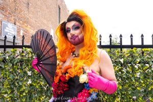 Performer with orange hair holding decorative fan poses against garden fence.