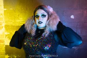 Drag performer in dark dress with colorful details poses against yellow and purple wall with dramatic lighting.