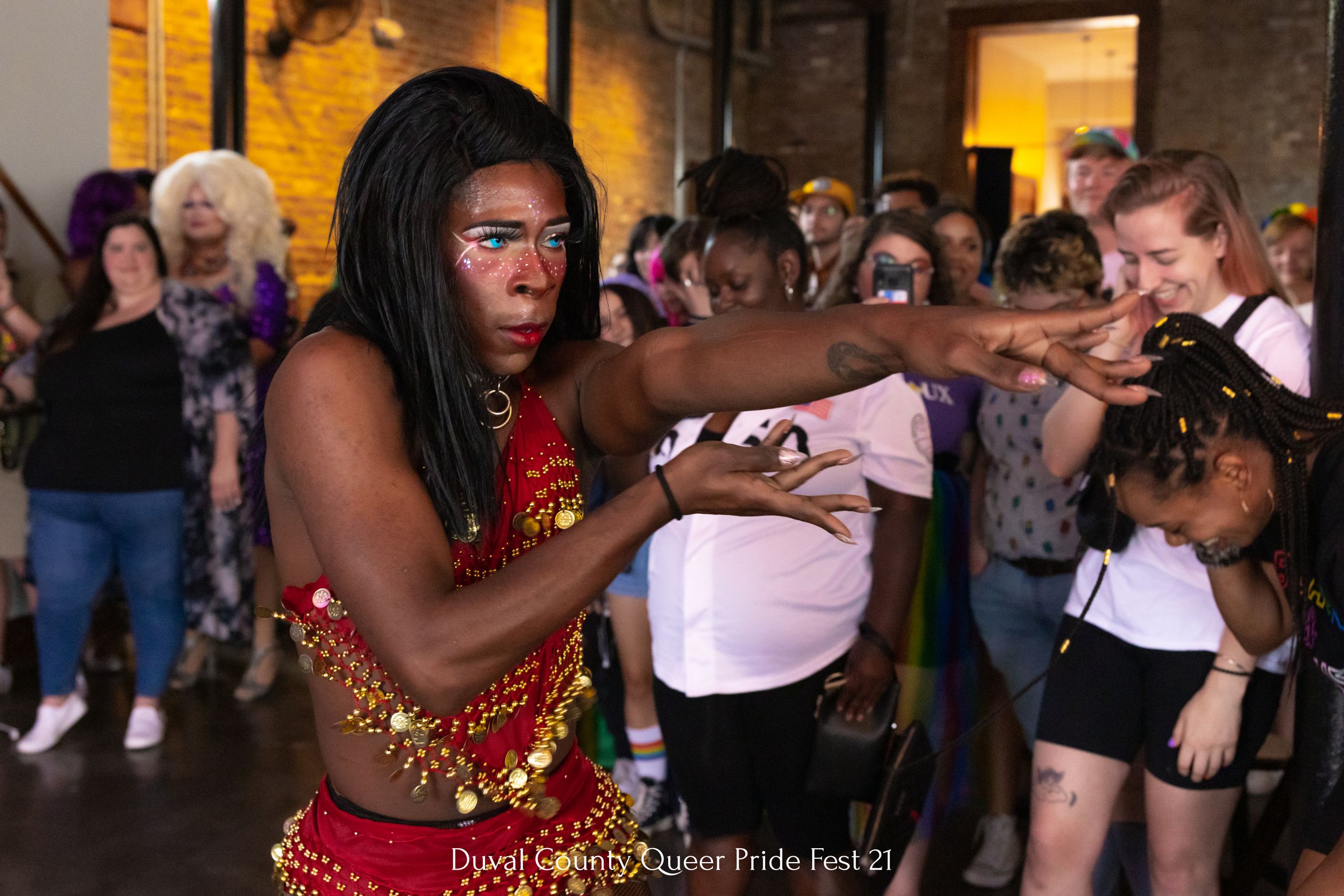Performer in red costume dances energetically during indoor pride celebration.