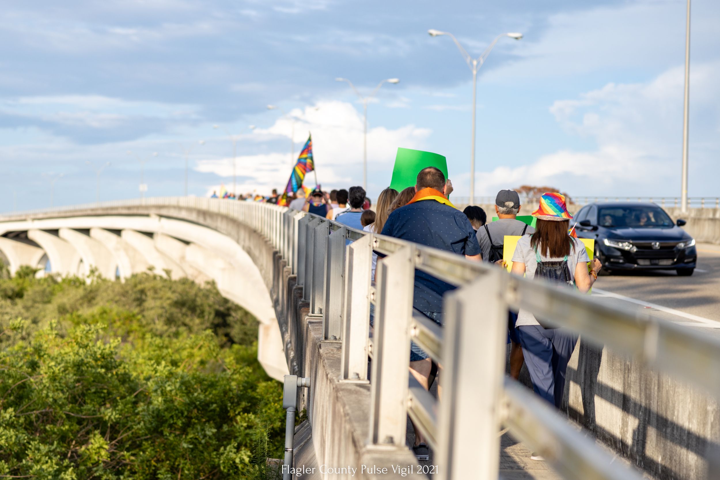 People gathering on a bridge with pride flags visible against cloudy sky.