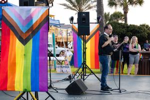 Rainbow pride flags frame an outdoor stage setup with speakers and performance equipment.