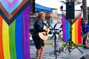 Performer with acoustic guitar plays between rainbow pride banners on outdoor stage.