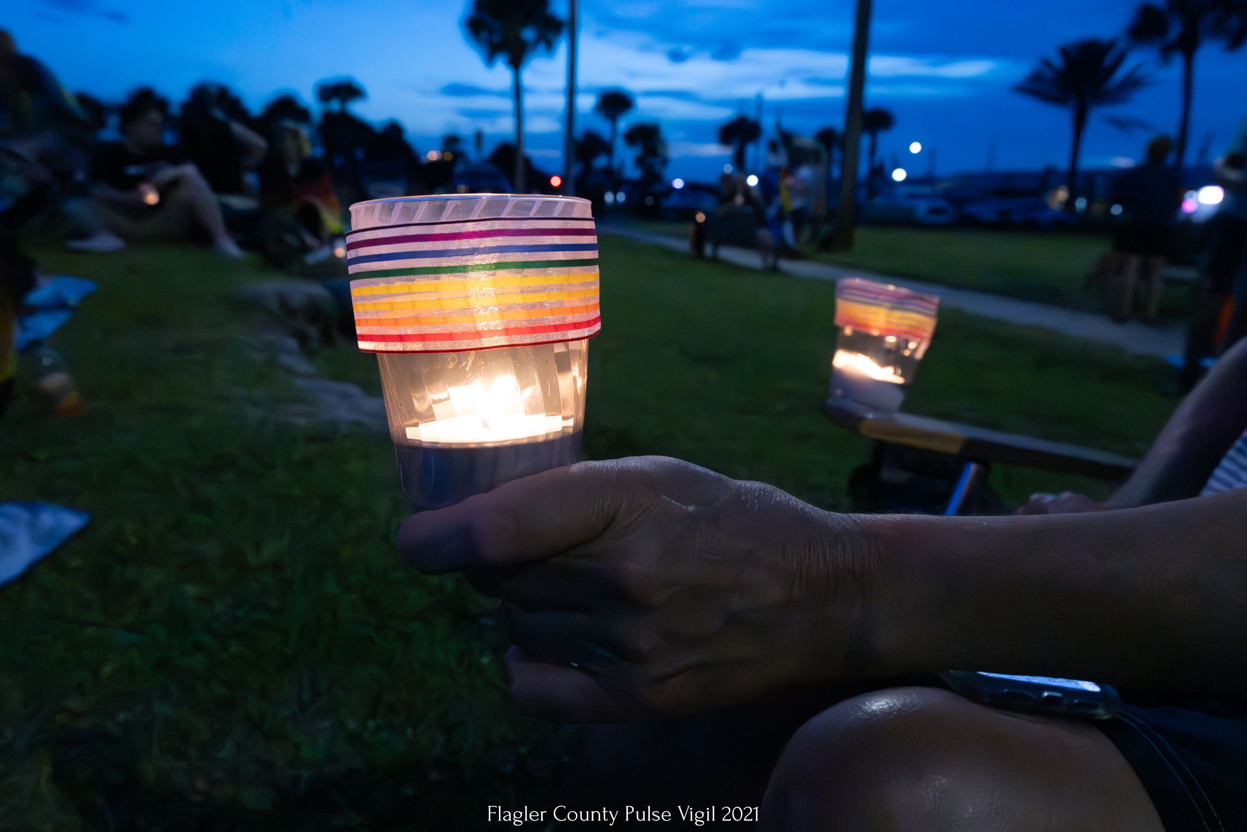 Hand holds illuminated candle holder at dusk with palm trees in background.