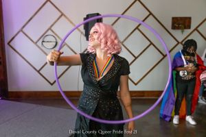 Performer with pink hair uses hula hoop on stage with geometric backdrop.