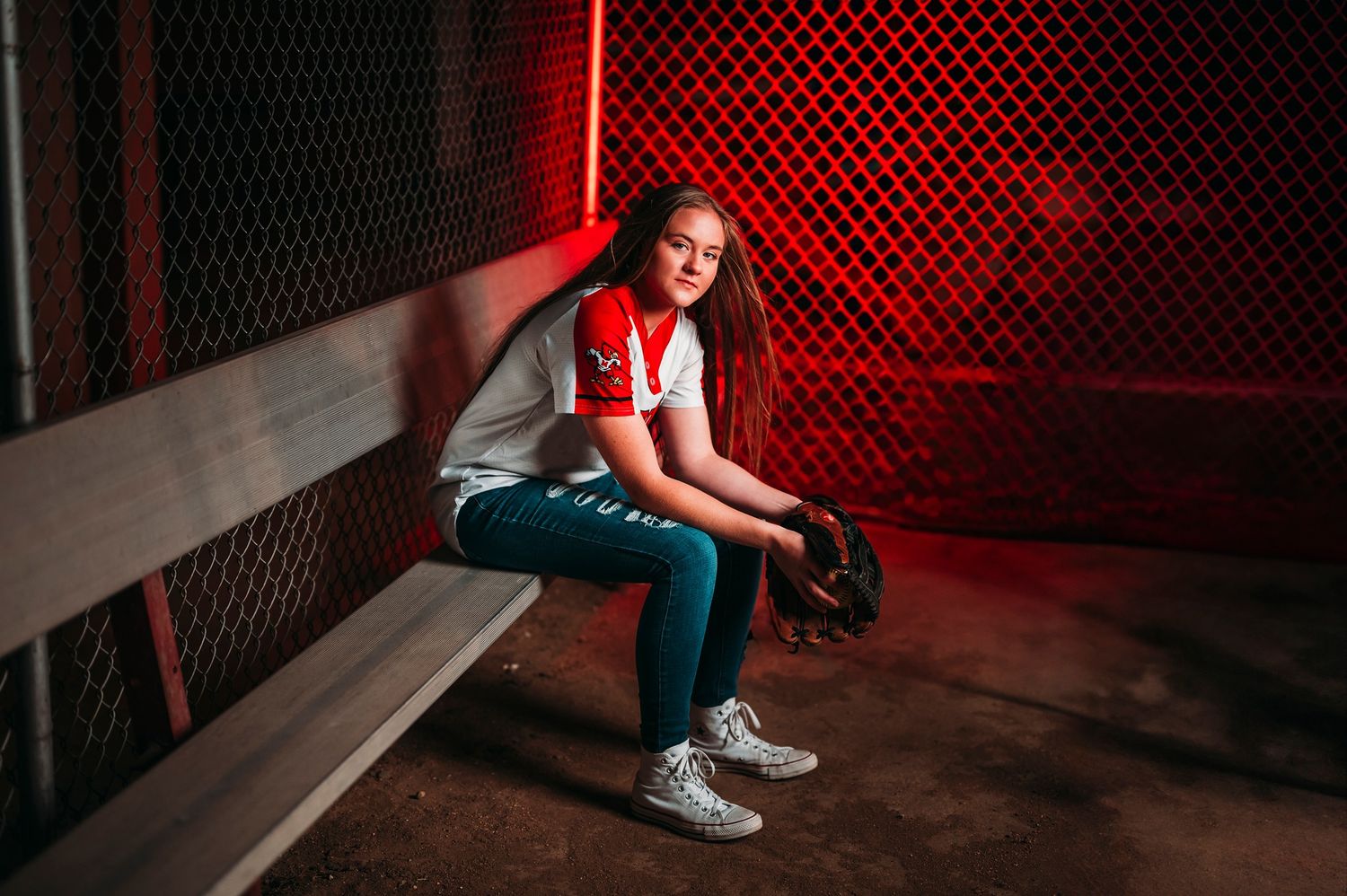 Teen girl softball player sitting on dugout bench with glove under moody red lighting.