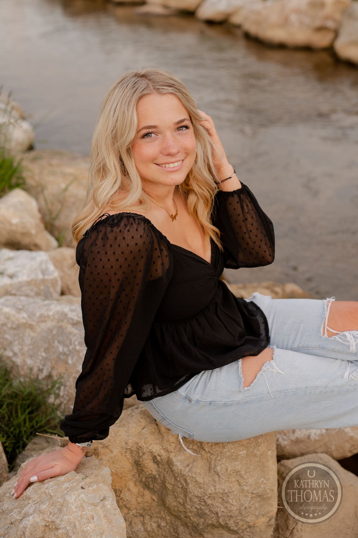 Outdoor portrait sitting on rocky stream bank in black sheer top and jeans.