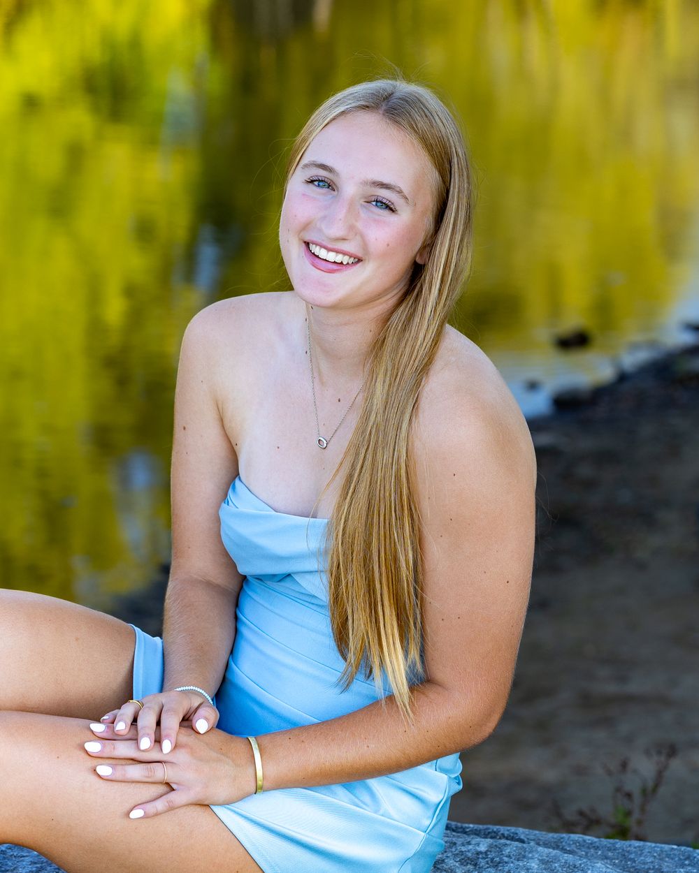 High school senior in a light blue dress sits outdoors with reflections of Concord River background on a sunny day for senior yearbook photo