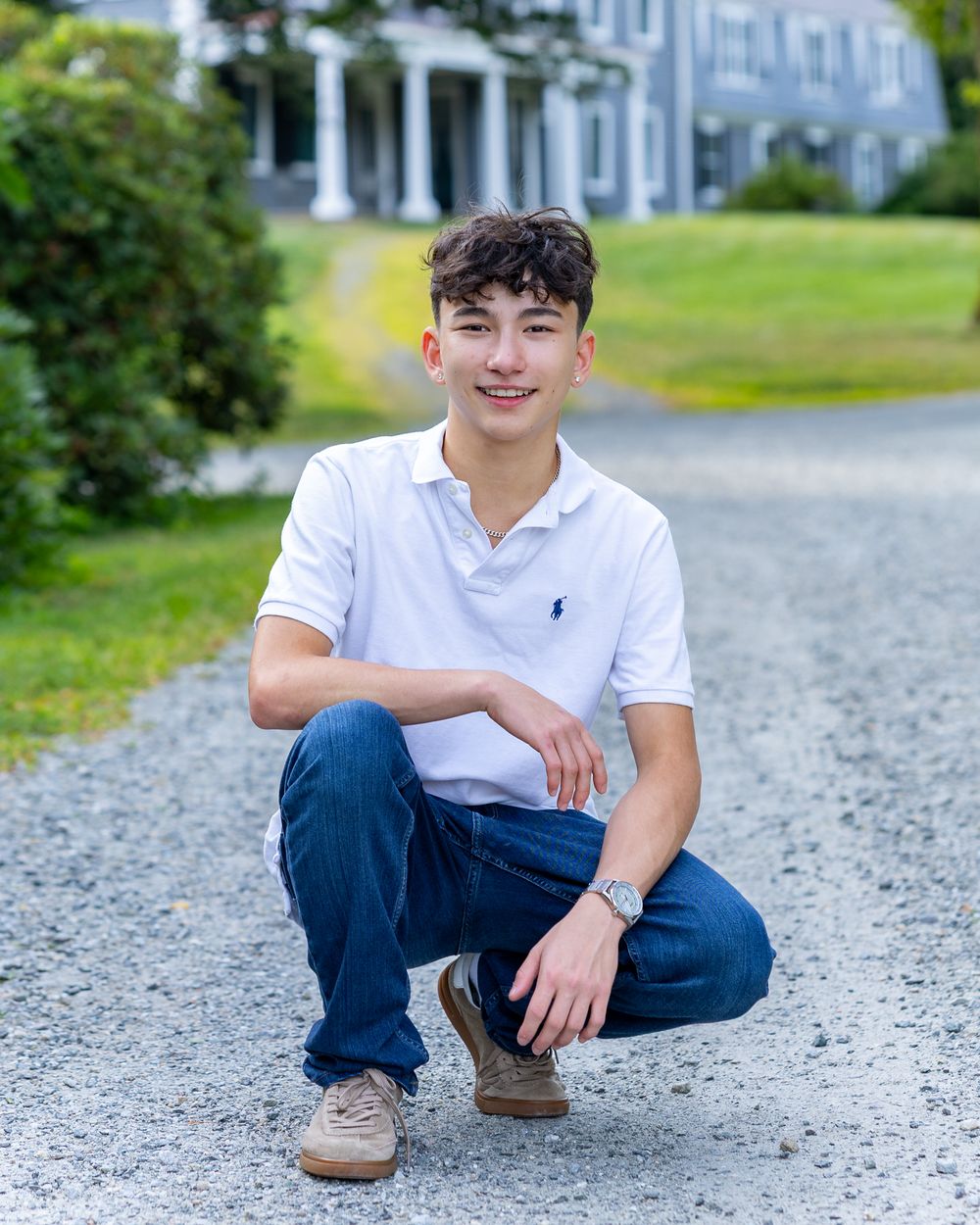 High school senior in a white polo shirt and jeans sits casually on a gravel path with an estate in the background during yearbook portraits