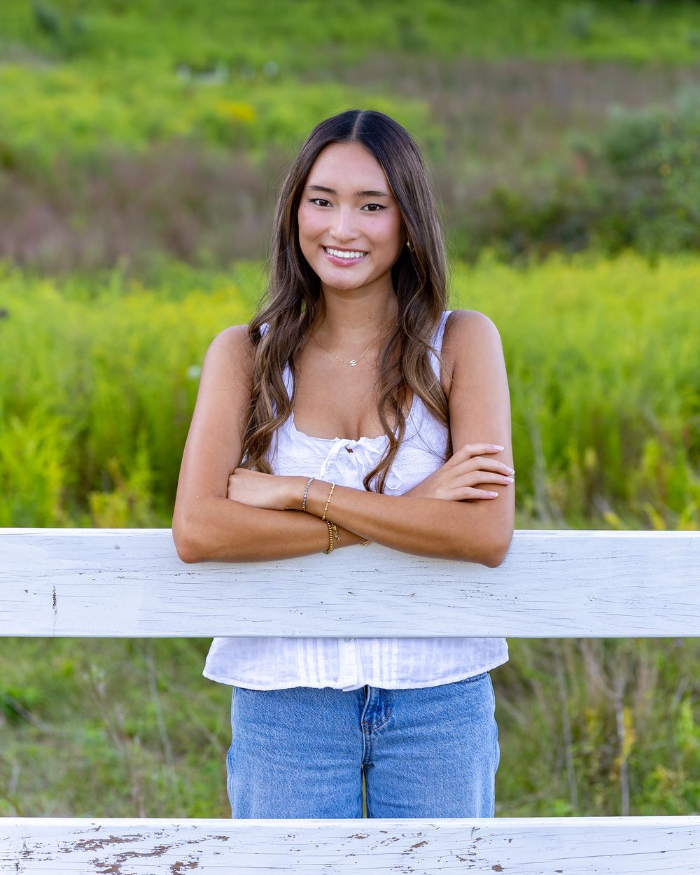 High school senior in a white sleeveless top and jeans leans against a wooden fence with green grass in the background for yearbook photos.