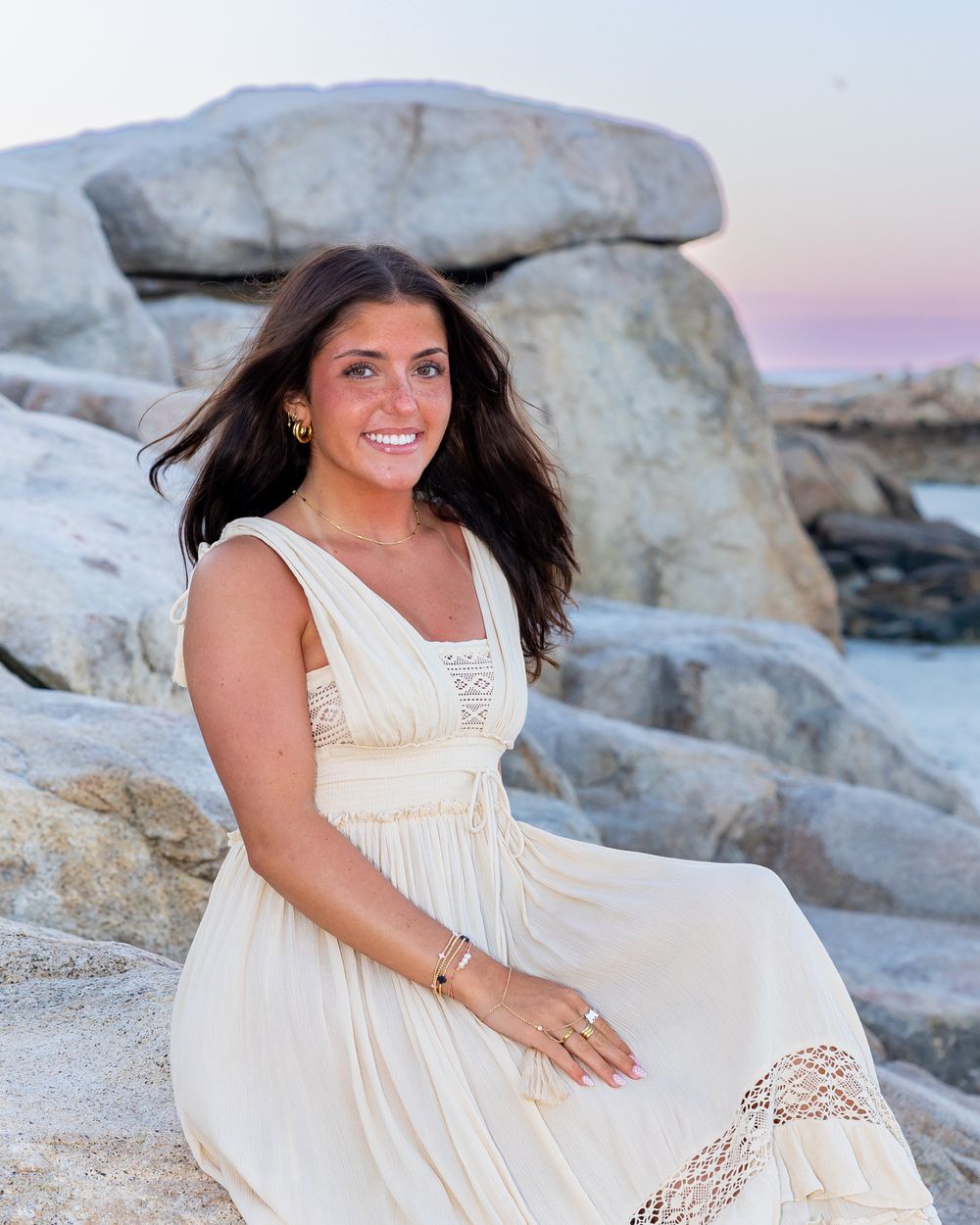 High school senior in flowing white dress poses in on rocks at beach during sunset photoshoot at Wingaersheek Beach Gloucester, MA