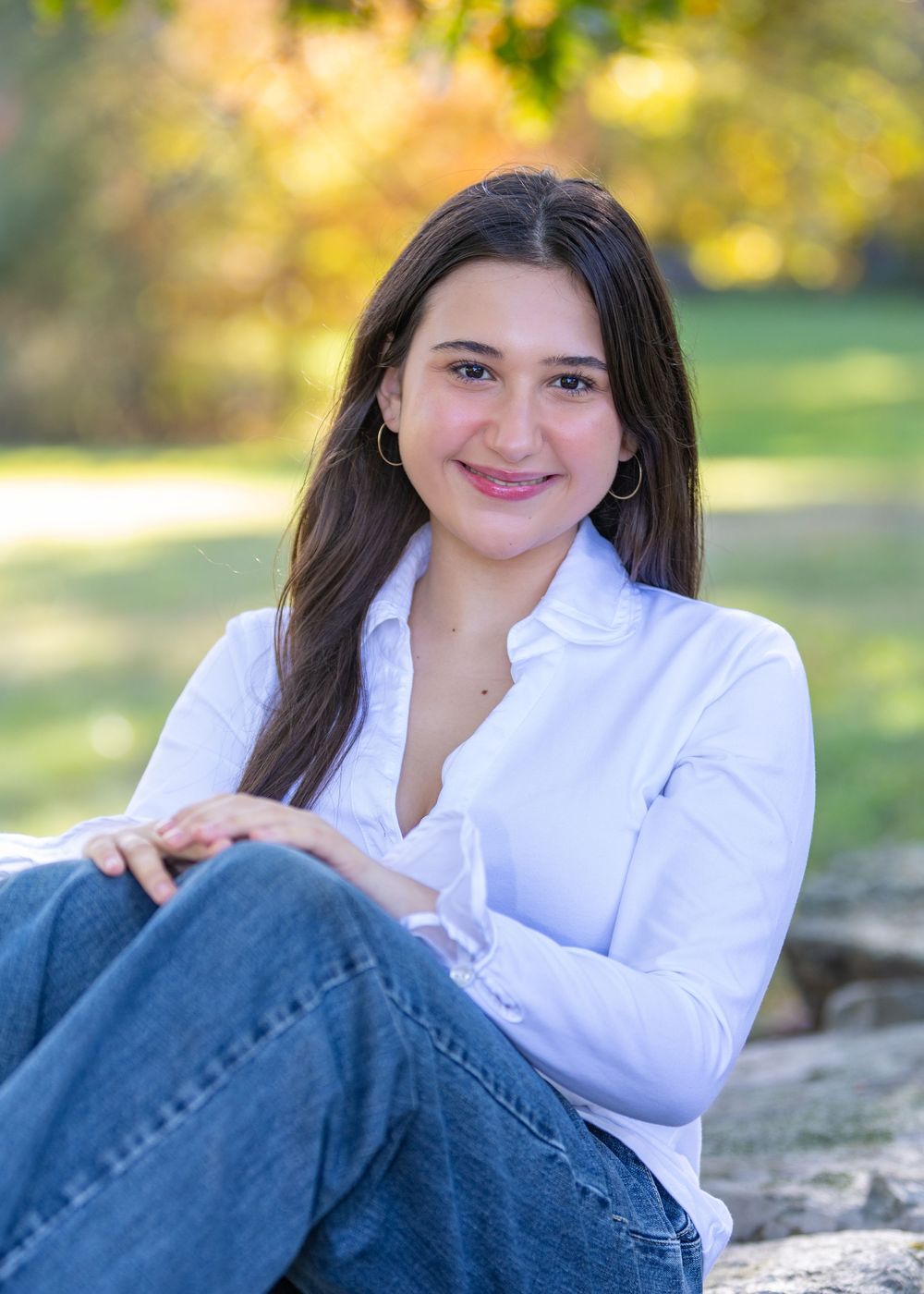 High school senior in a white button-down shirt & jeans sits casually on a stone wall outdoors on a sunny autumn day during senior portraits
