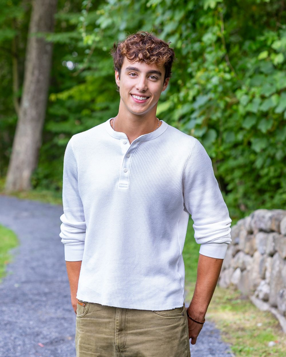 High school senior in a white henley shirt & khaki pants stands on a gravel path with green trees in the background during senior portraits