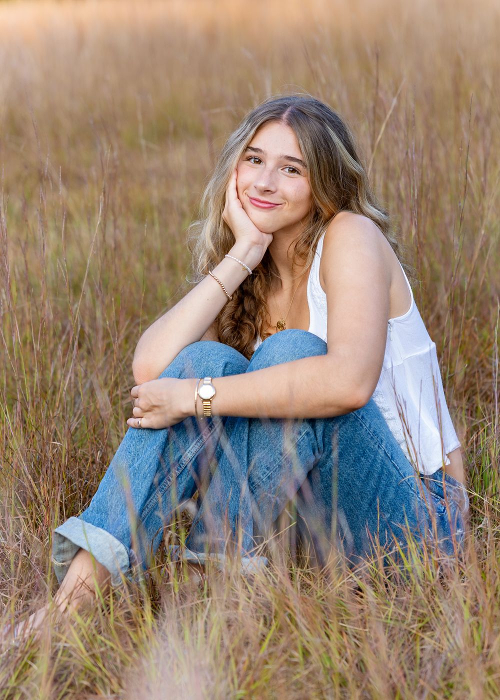 A high school senior in a white top and jeans sits casually in a field of tall brown grass during golden hour during a photo session Sudbury