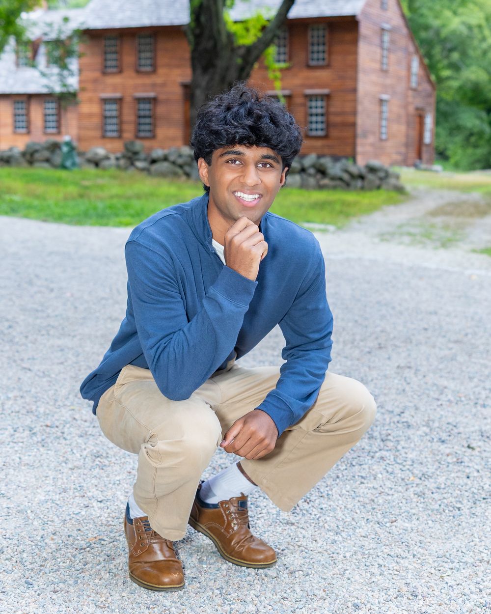 A high school senior in a blue quarter-zip pullover & khaki pants crouches on a gravel path with in front of  Hartwell Tavern in Lincoln, MA