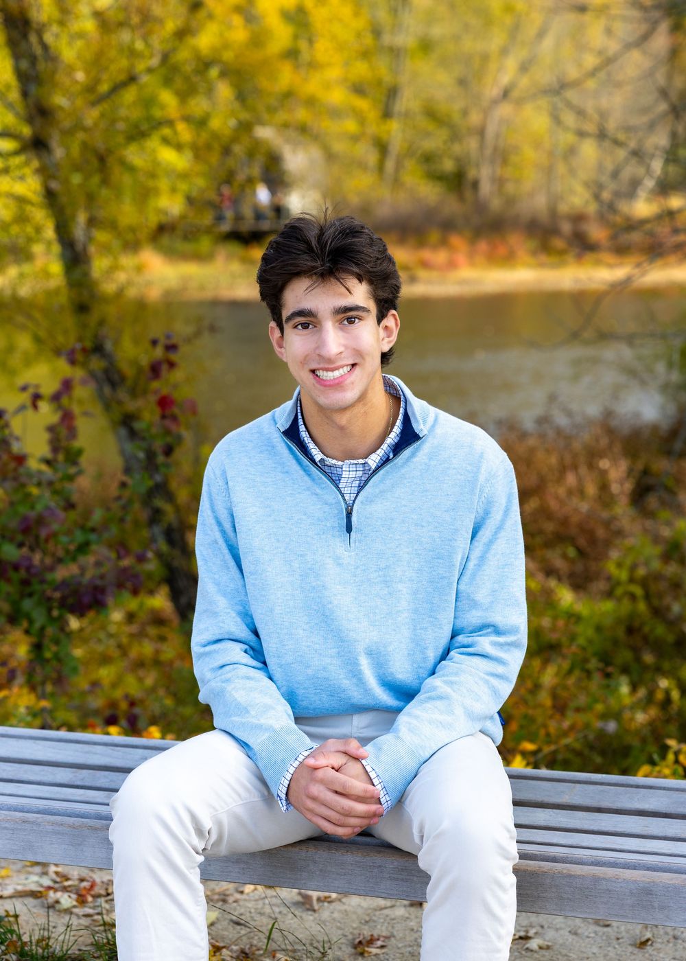 High school senior in a light blue quarter-zip sweater & khaki pants poses against a backdrop of autumn foliage at North Bridge Concord, MA