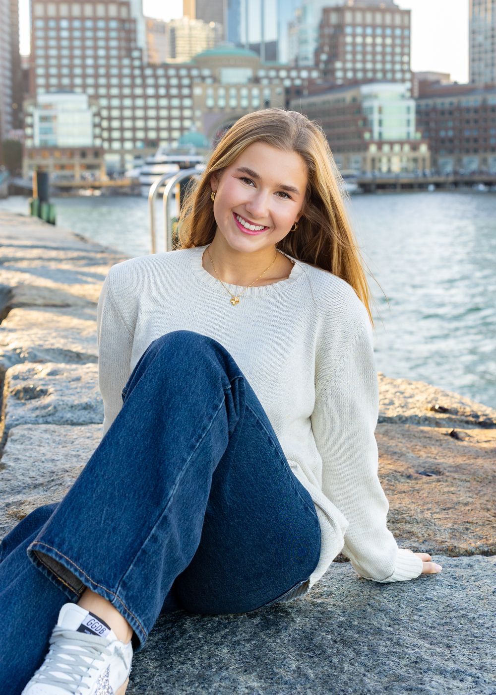 High school senior in a white sweater and jeans sits casually in Boston's Seaport with city buildings in the background.