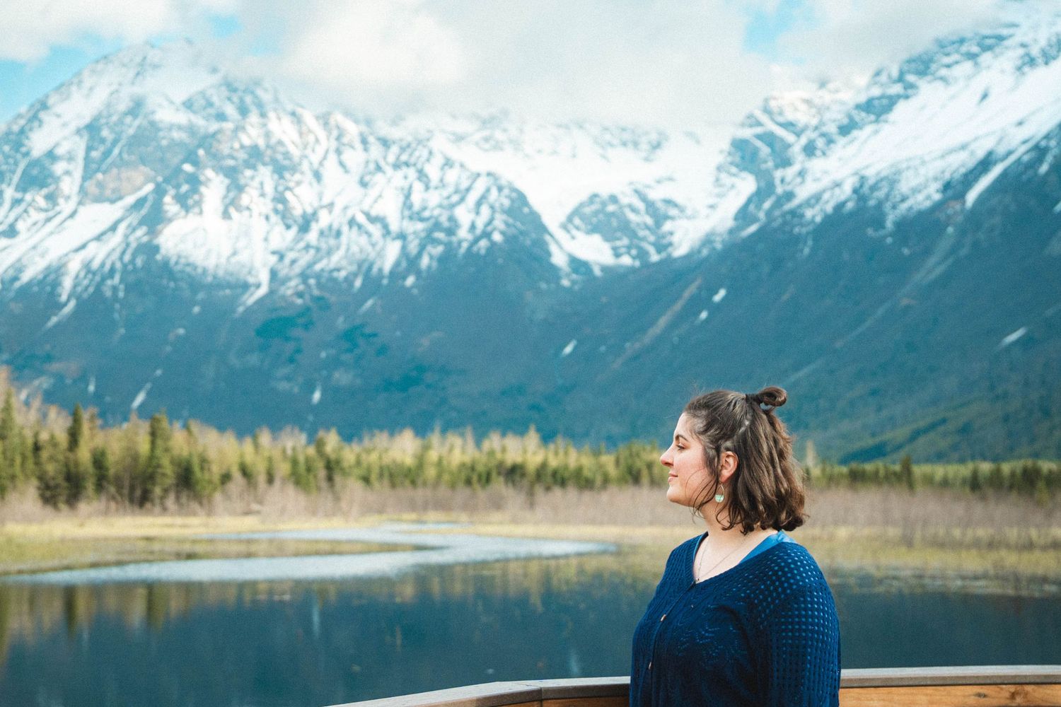A young woman with long dark hair and blue eyes smiles softly outdoors wearing a teal top and leaf earring.
