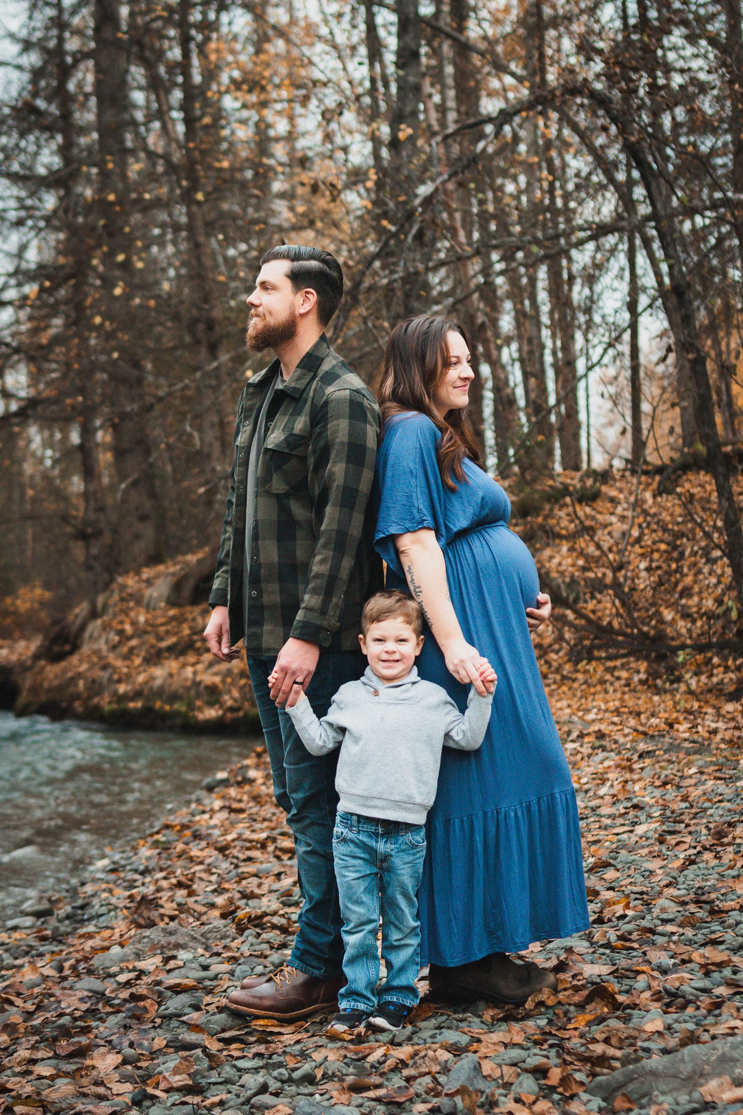 Pregnant woman in blue dress and bearded man hold hands with toddler boy outdoors in autumn forest setting.
